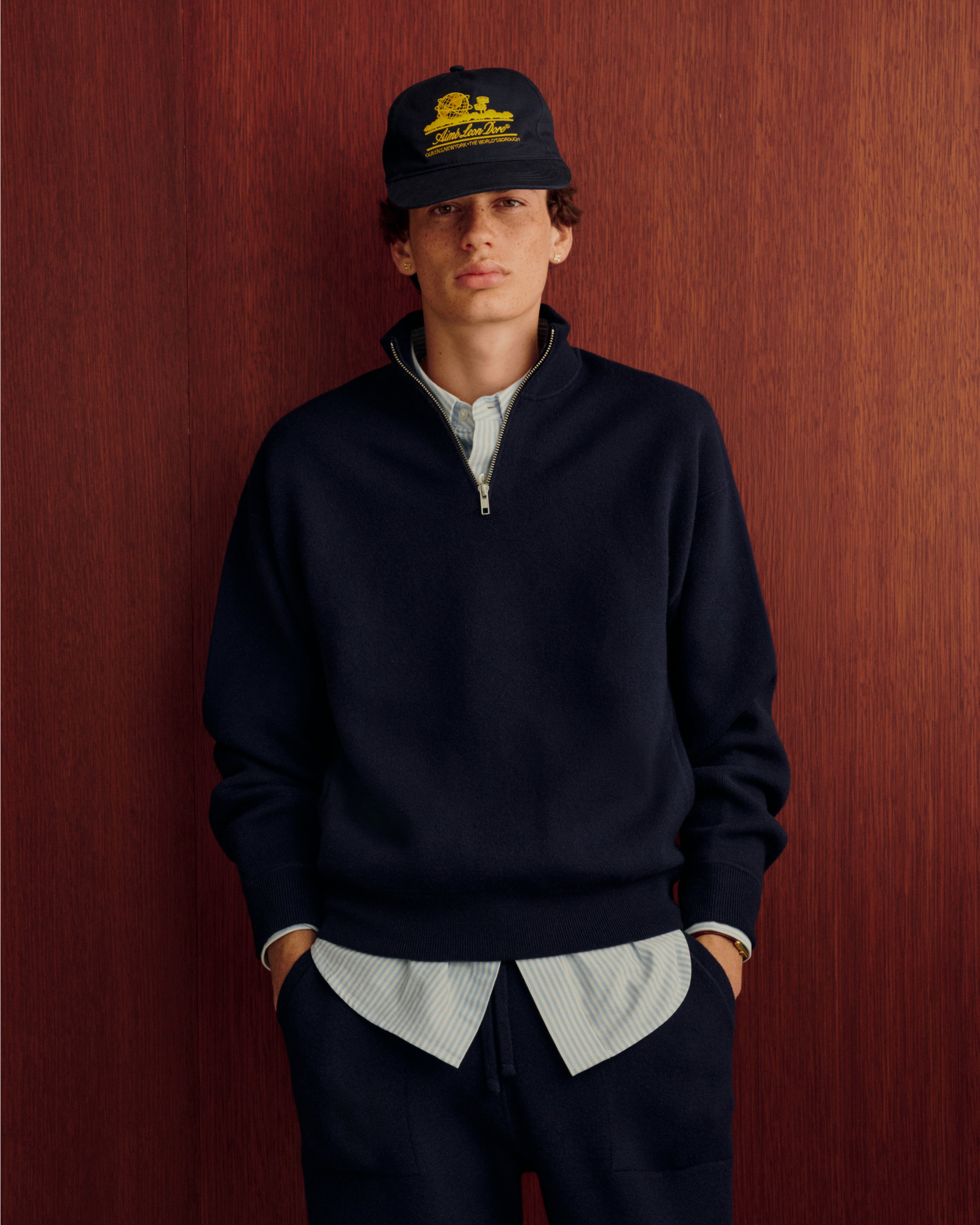 Young man in the Quarter Zip Pavilion Cashmere Pullover stands against a red wooden wall with hands in pockets.