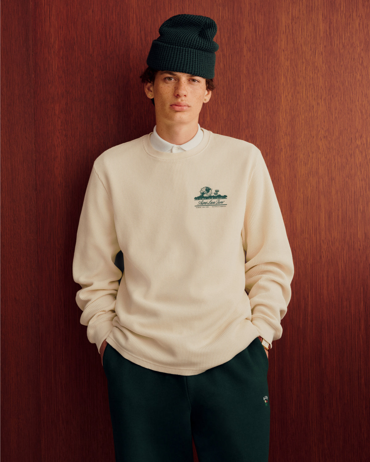 Person wears Long-Sleeve Unisphere Waffle Thermal, white shirt, green pants, and beanie in front of a wooden wall.