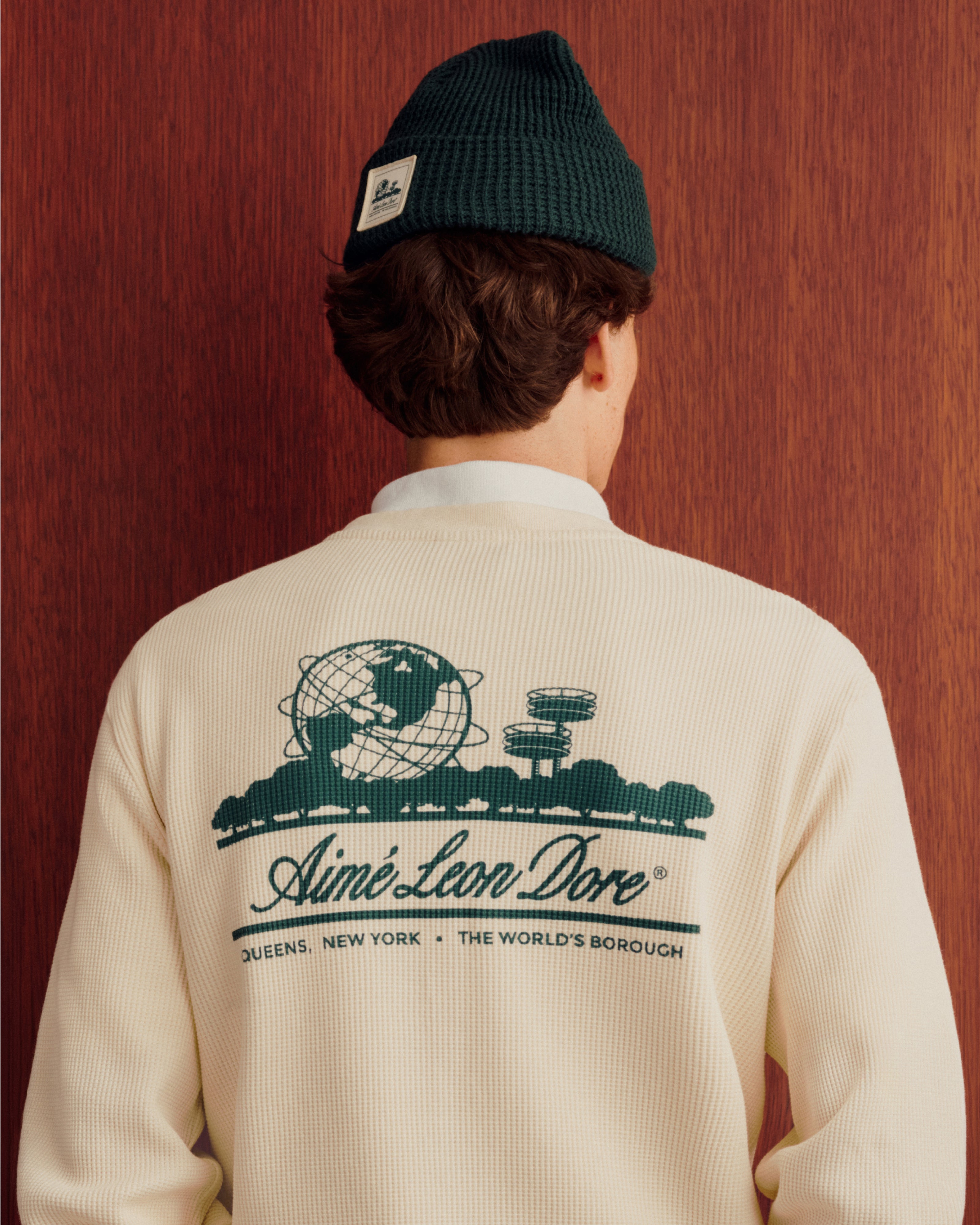Person wearing Waffle Knit Beanie and a cream sweater with green globe graphic and Aimé Leon Dore text on back.