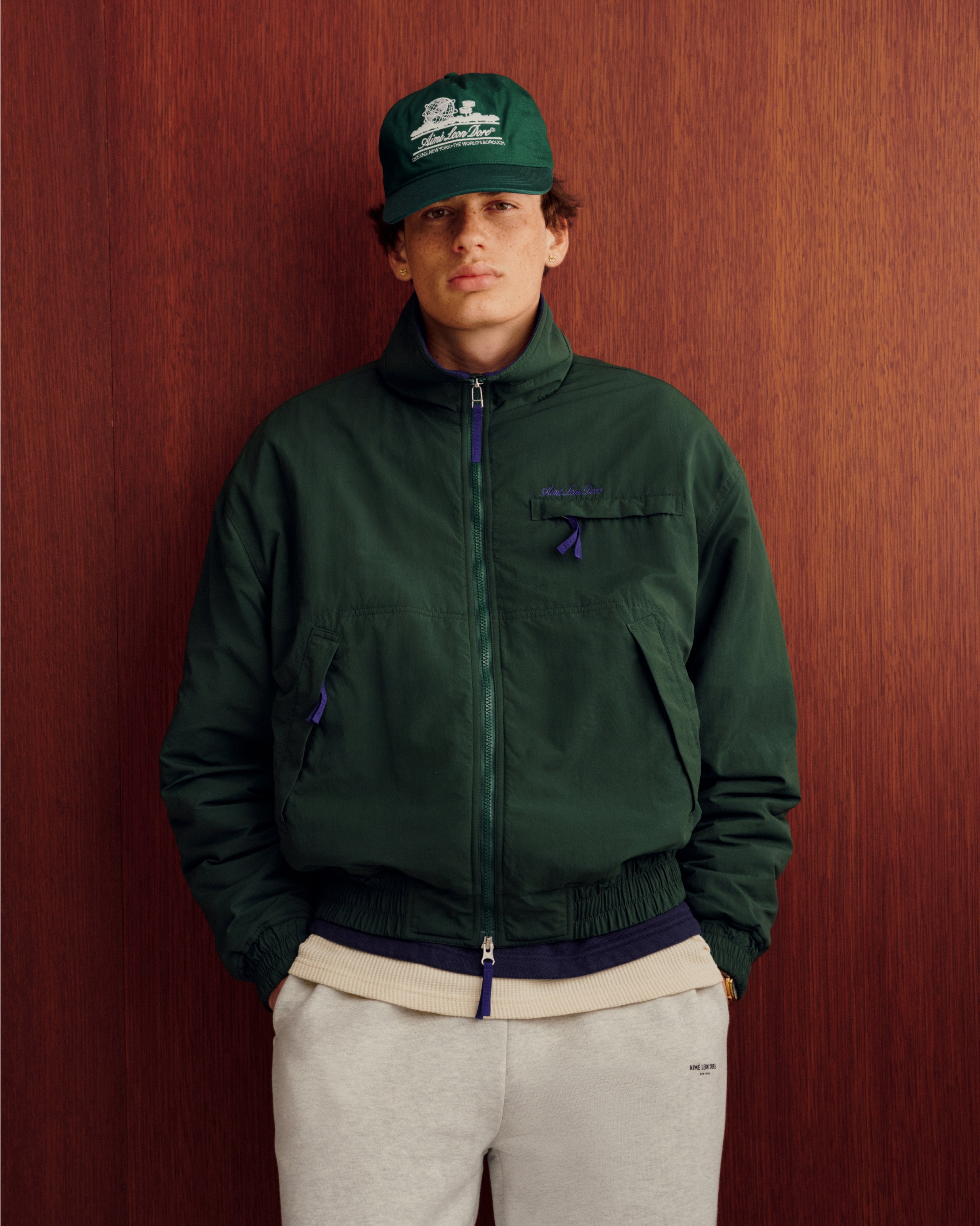 Person wears the Unisphere Hat and green jacket, hands in gray sweatpants pockets, standing before a wooden wall.
