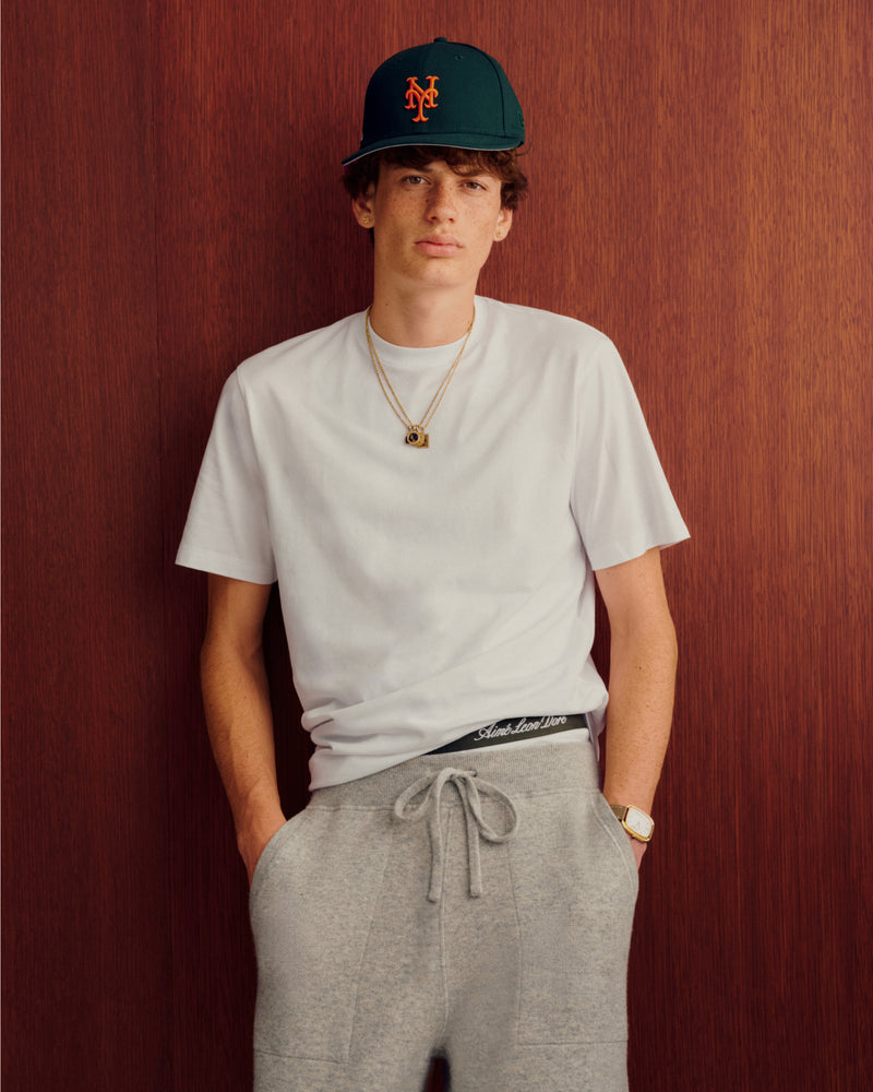 Young man in white tee, gray sweats, and ALD/New Era Mets Ballpark Hat stands by a wooden background.