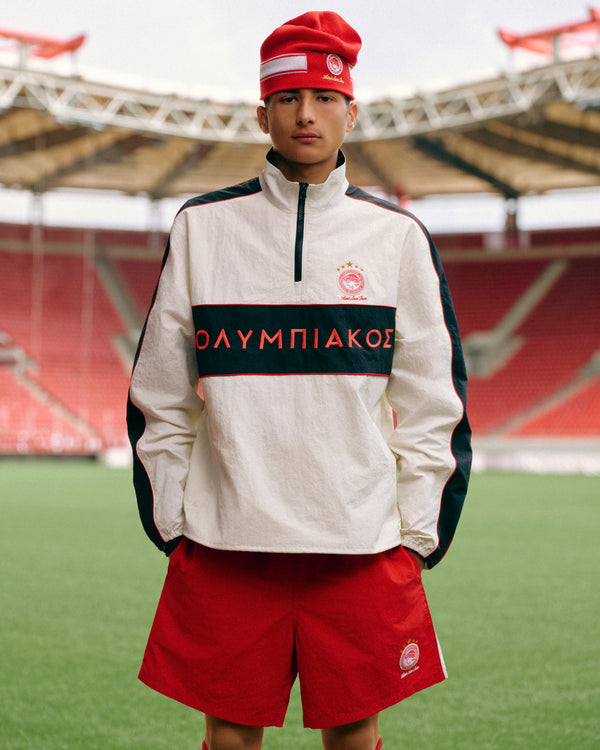 Person wearing an ALD / Olympiacos F.C. Nylon Quarter Zip Pullover with red shorts and a red beanie on a soccer field.