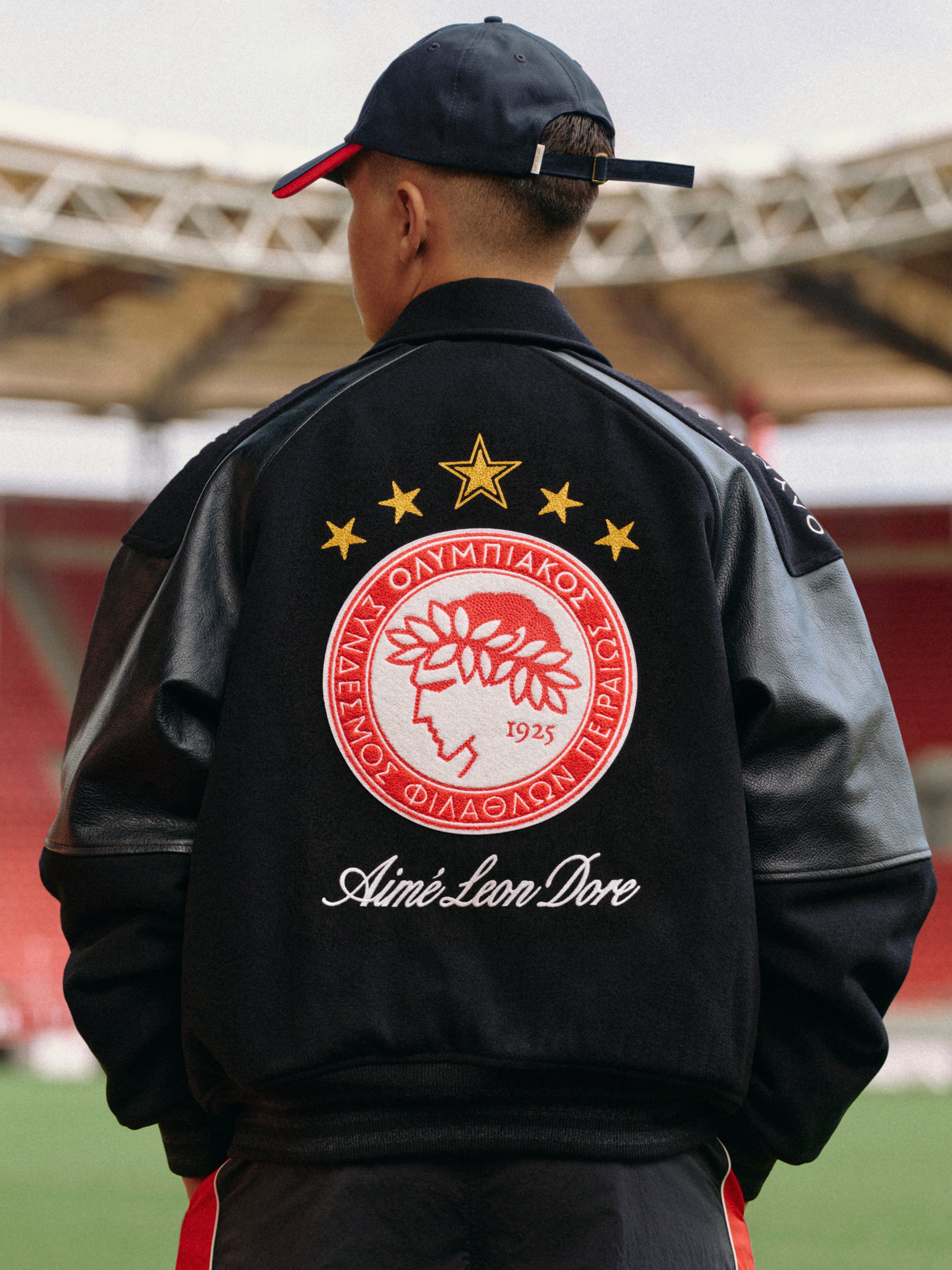 A person wears an ALD / Olympiacos F.C. Team Leather Jacket with a large team emblem and script text on the back.