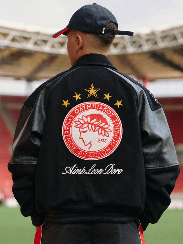 A person wears an ALD / Olympiacos F.C. Team Leather Jacket with a large team emblem and script text on the back.