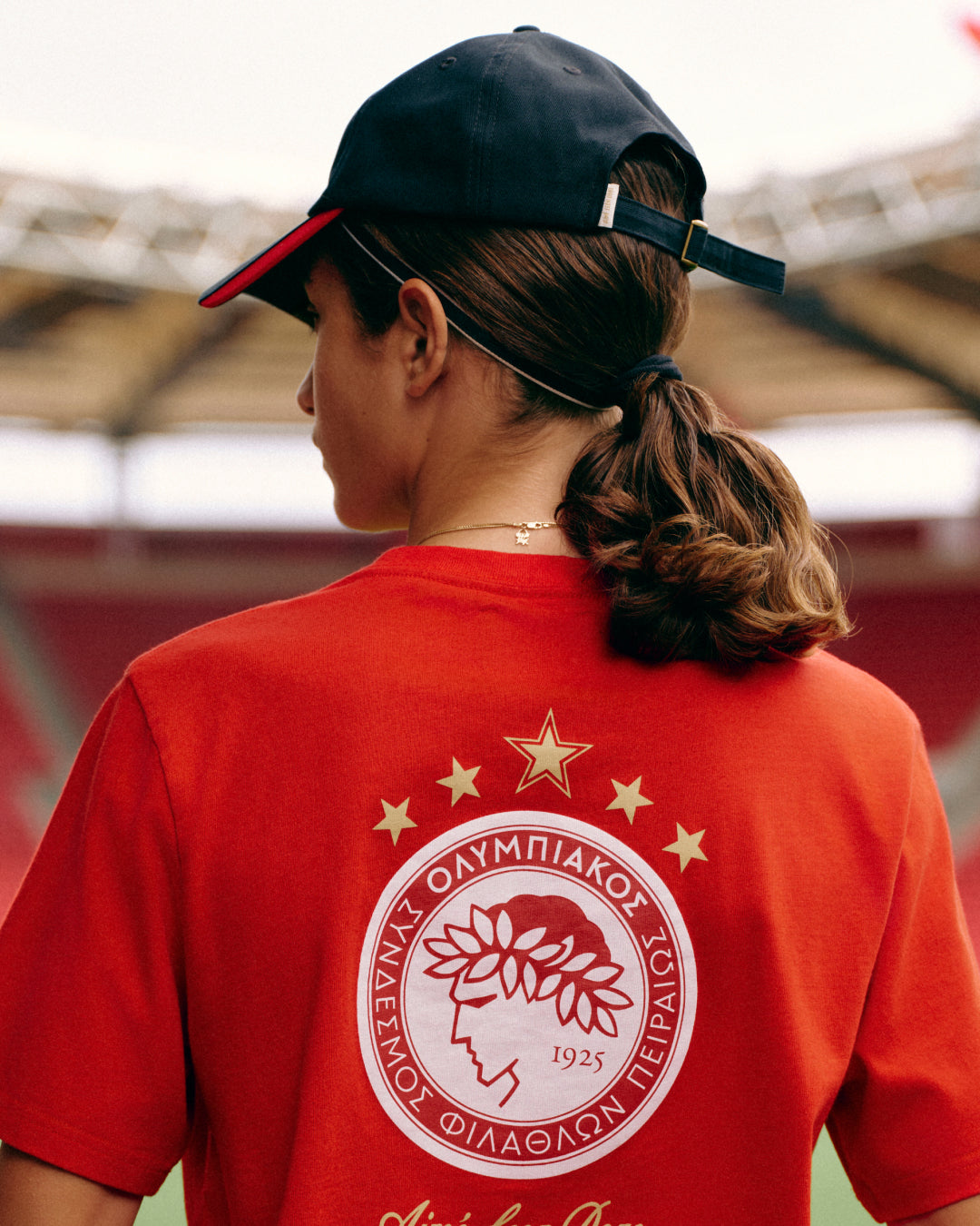 Person wearing a red ALD / Olympiacos F.C. Souvenir Tee featuring a circular team crest and gold stars on the back.