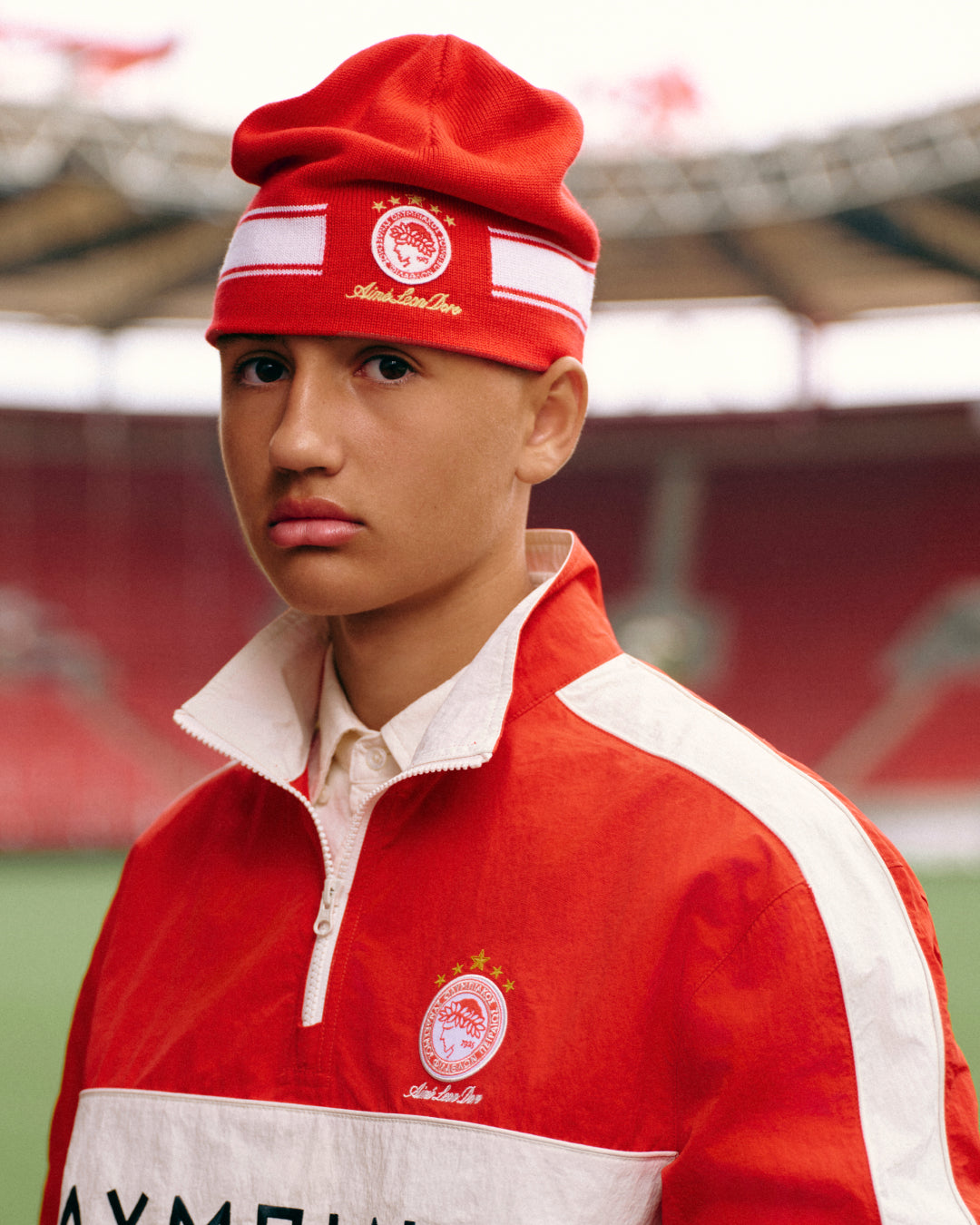 A person wears a red ALD / Olympiacos F.C. Stadium Beanie with white stripes and a soccer club emblem in a stadium.