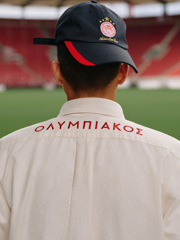 Person wearing an ALD / Olympiacos F.C. Logo Oxford Shirt with red Greek lettering and a dark baseball cap in a stadium.