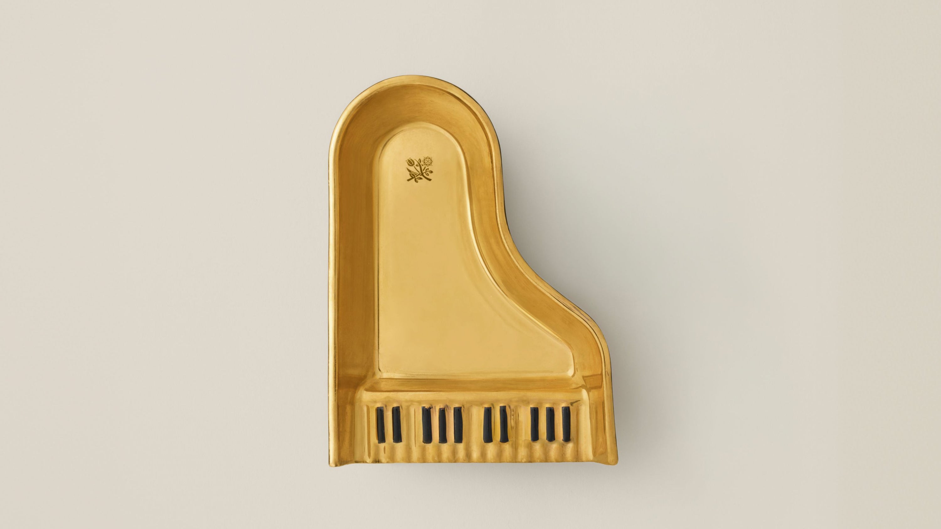 A gold-finished ceramic grand piano ashtray , featuring black key accents at the bottom, shown against a neutral light beige background.