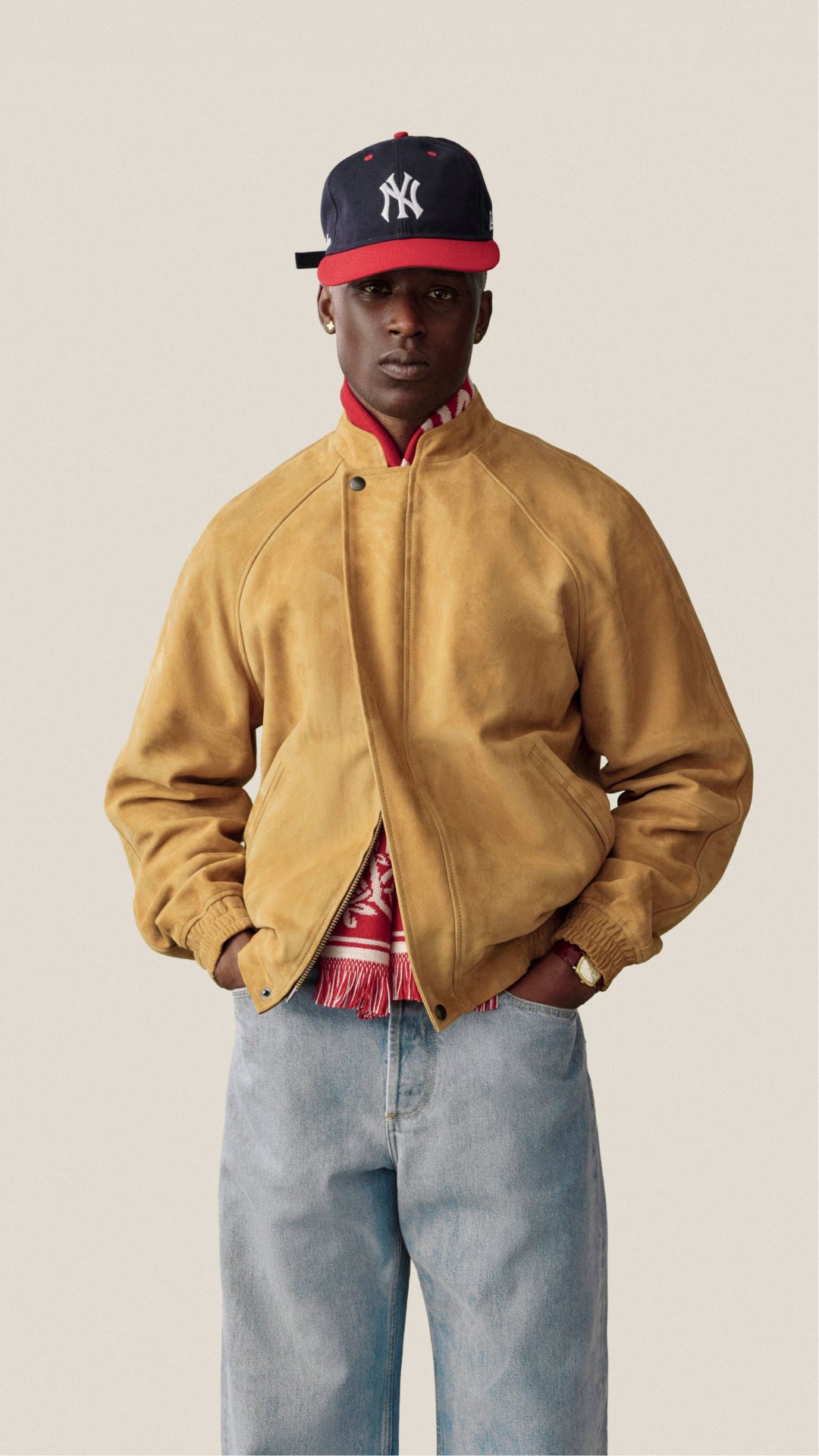A model wearing a tan suede baseball jacket, a navy and red New York Yankees cap, a red patterned shirt, and light blue jeans, standing against a solid light beige background.