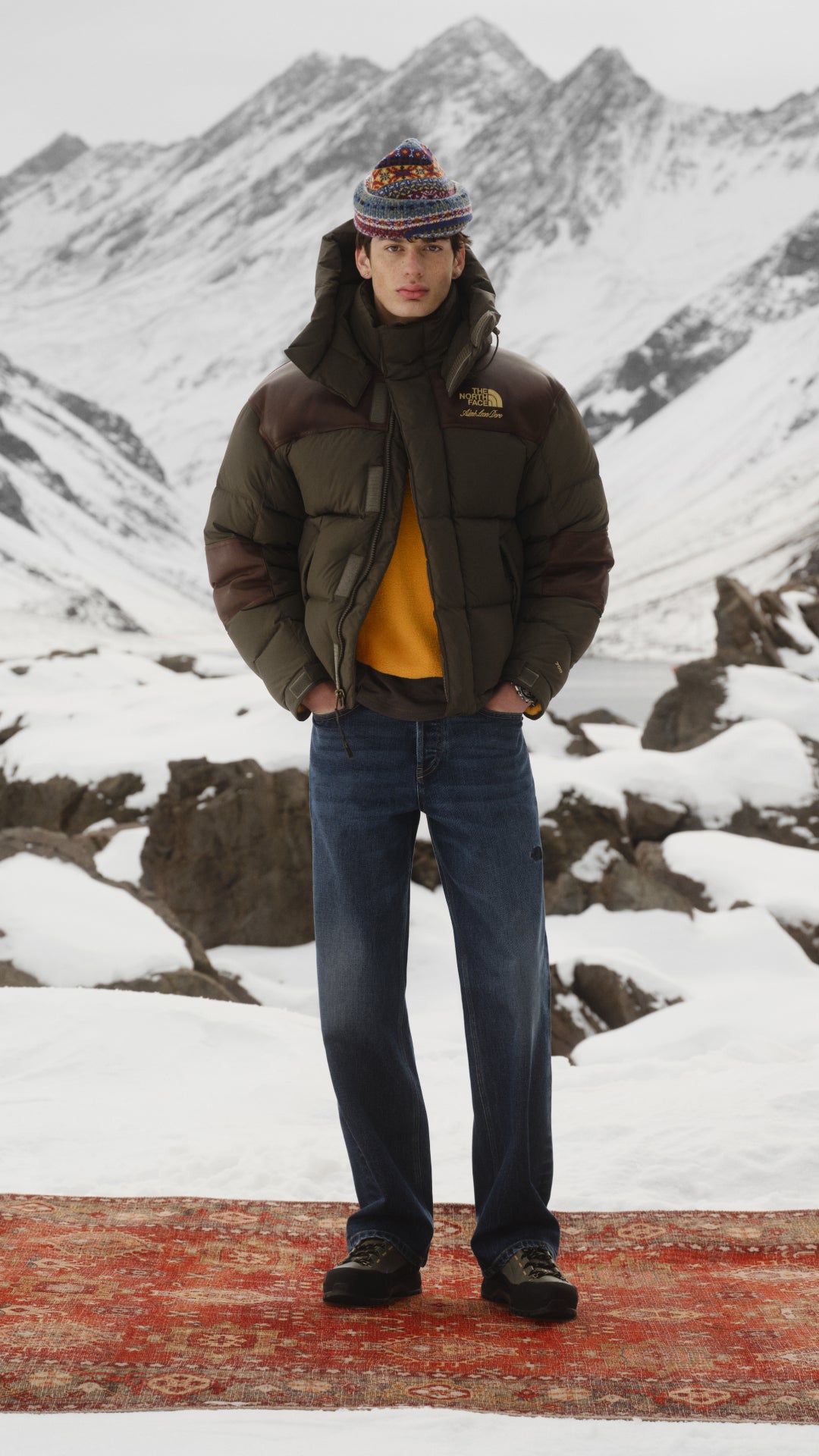 man standing in front of snowy mountain in green puffer jacket and jeans