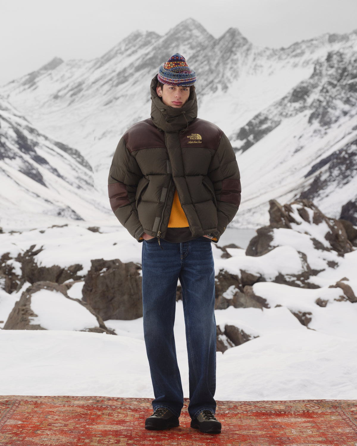 A person in the ALD / The North Face Baltoro Jacket stands on a rug in snowy mountains with rocky peaks behind.