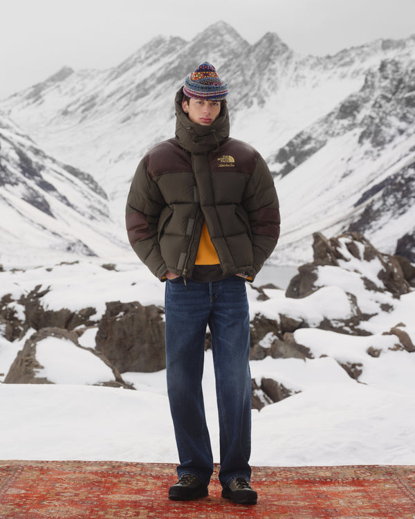 A person in the ALD / The North Face Baltoro Jacket stands on a rug in snowy mountains with rocky peaks behind.