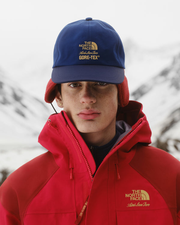 Person in red North Face jacket and ALD / The North Face GORE-TEX Hat stands outdoors with snowy mountains behind.