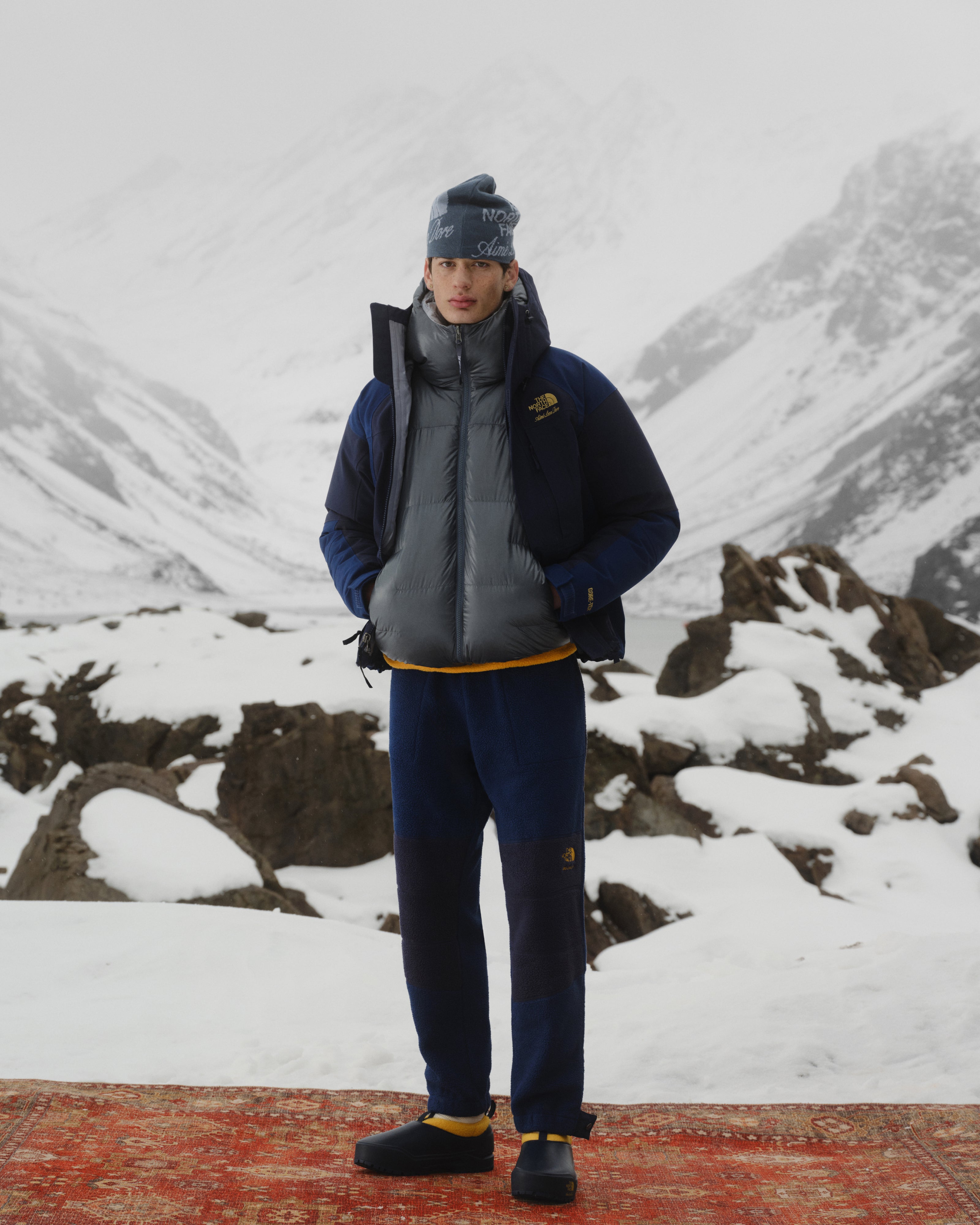 A person in the ALD/The North Face GORE-TEX 2000 Mountain Jacket stands on a red rug in snowy mountains with rocks.