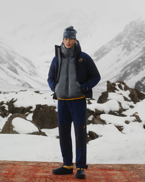 Person in winter clothing wears ALD/The North Face Base Camp Mule on a red rug in a snowy, mountainous landscape.