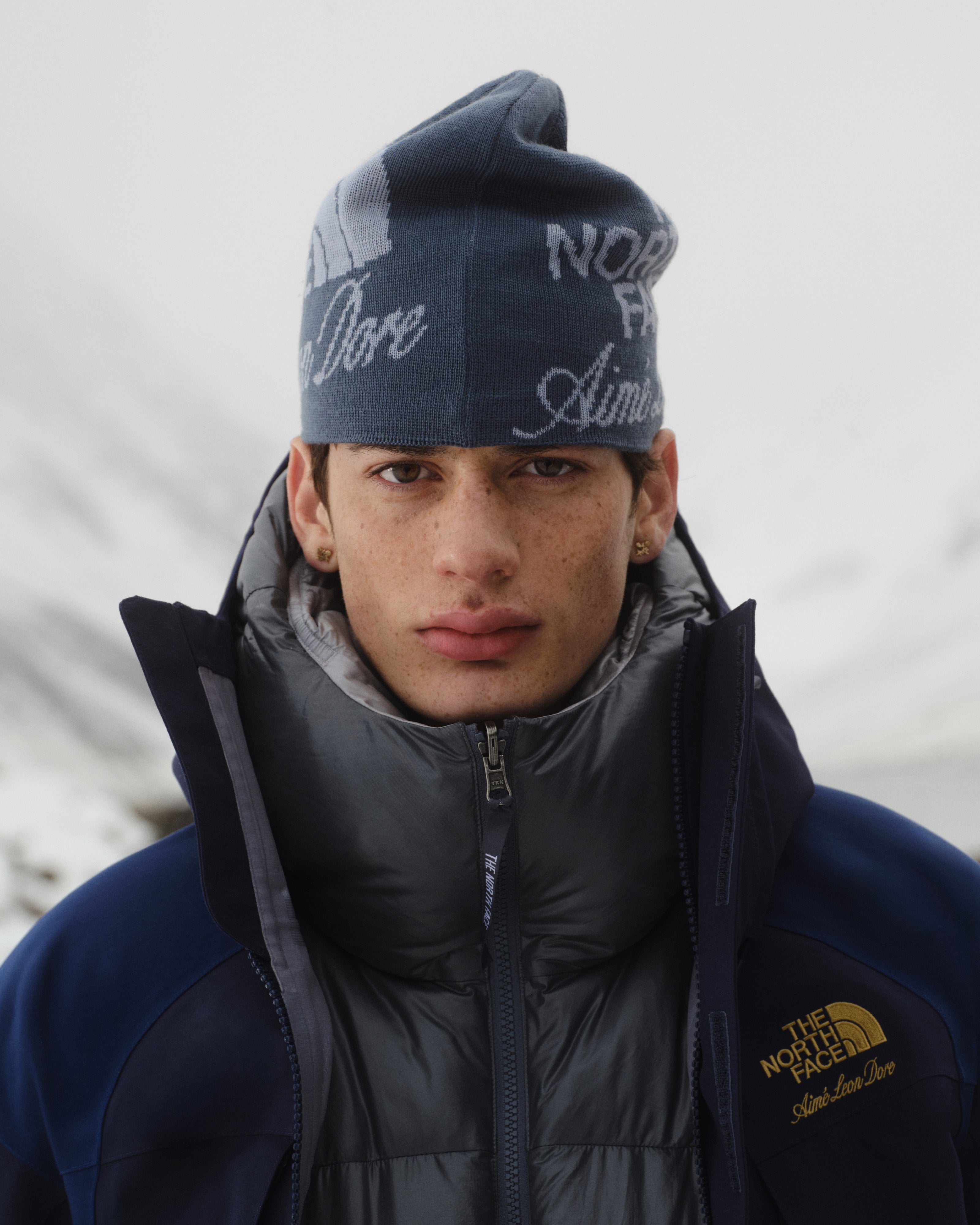 A young man wears an ALD / The North Face Jacquard Beanie outdoors with snowy mountains in the background.