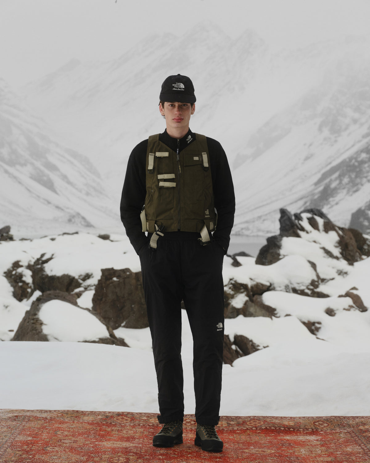 Person wearing the ALD / The North Face Nylon Logo Hat stands on a red rug in a snowy mountain landscape.
