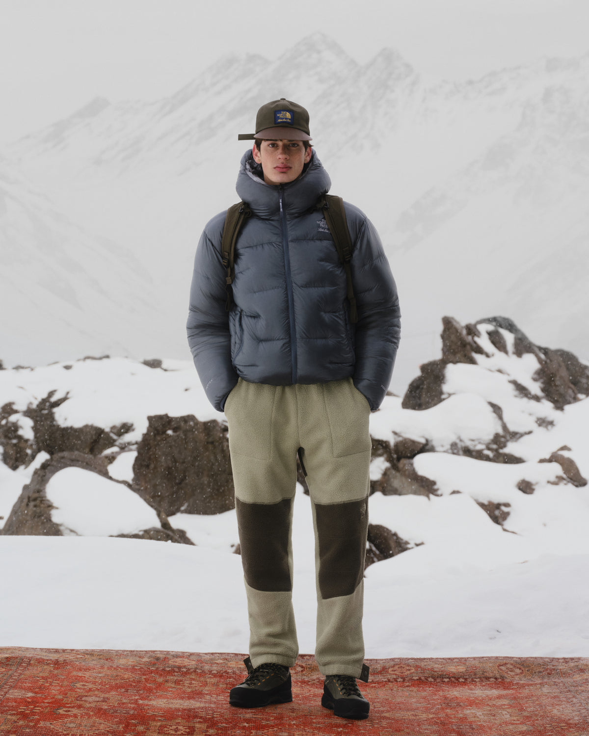Someone in winter gear wears the ALD / The North Face Leather Brim Logo Hat, snowy mountains behind them.