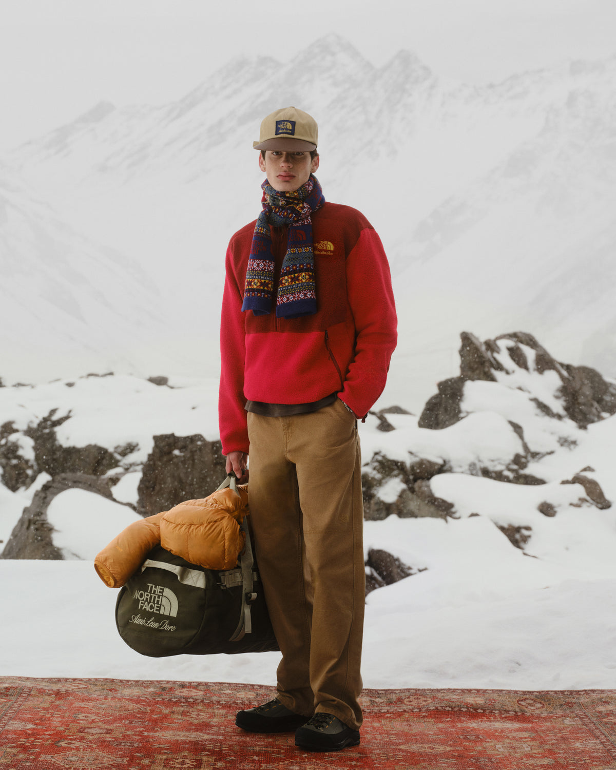 A man in winter clothes wears the ALD / The North Face Leather Brim Logo Hat on a snowy mountain, holding a duffel bag.