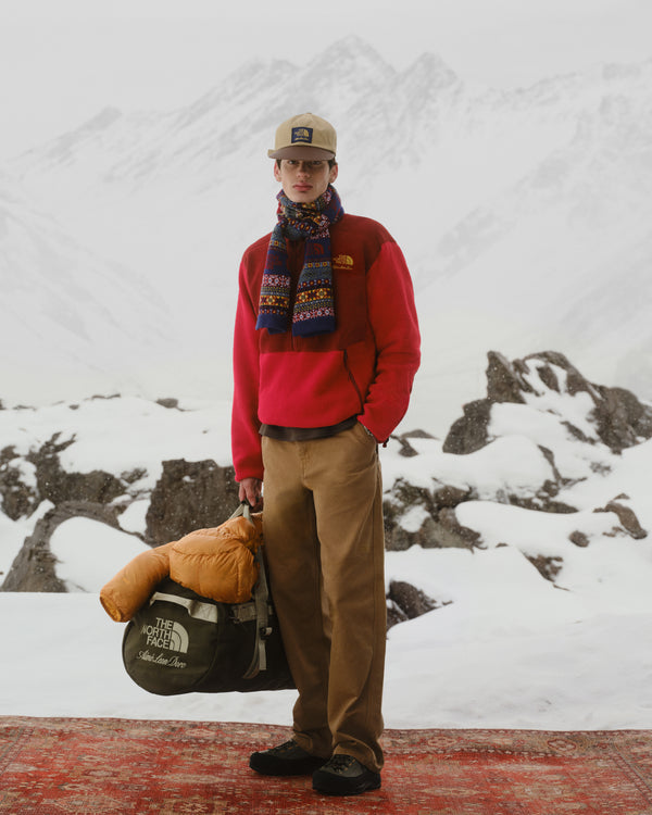 A man in winter clothes wears the ALD / The North Face Leather Brim Logo Hat on a snowy mountain, holding a duffel bag.