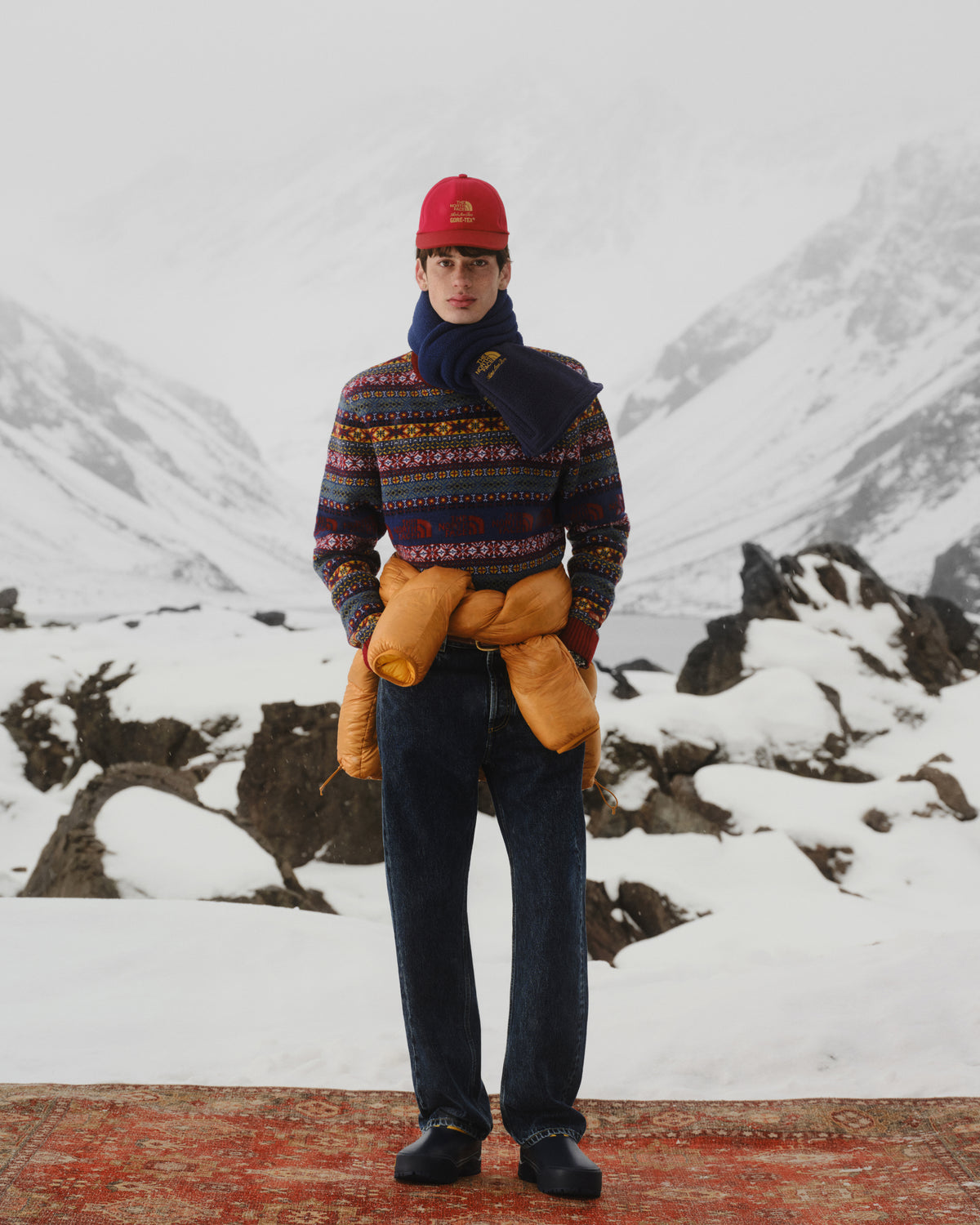 A person in winter clothes and a red hat stands on a snowy rug, wearing the ALD / The North Face Denali Fleece Scarf.