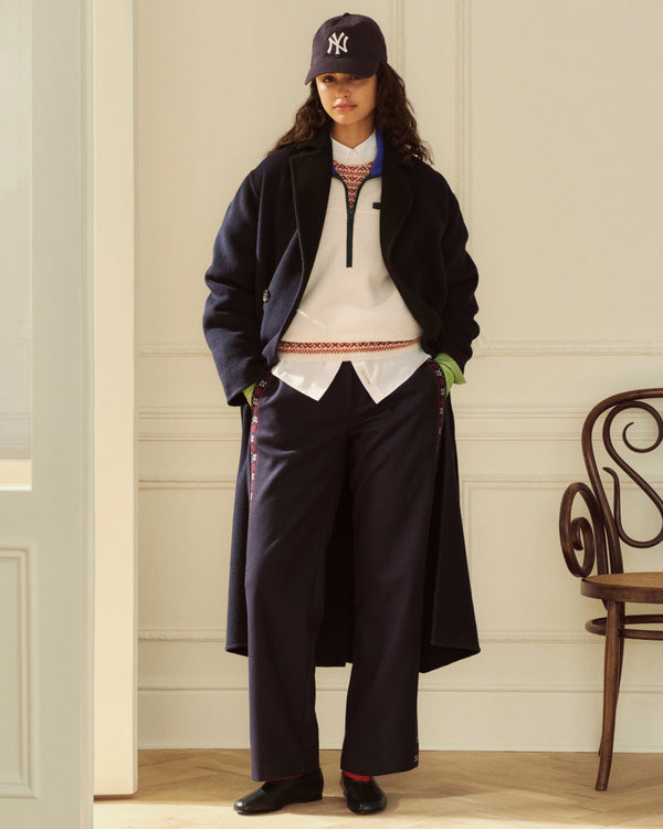 A model wears a dark coat over a Button Down Poplin Shirt, a patterned zip-up pullover, dark trousers, and a baseball cap.