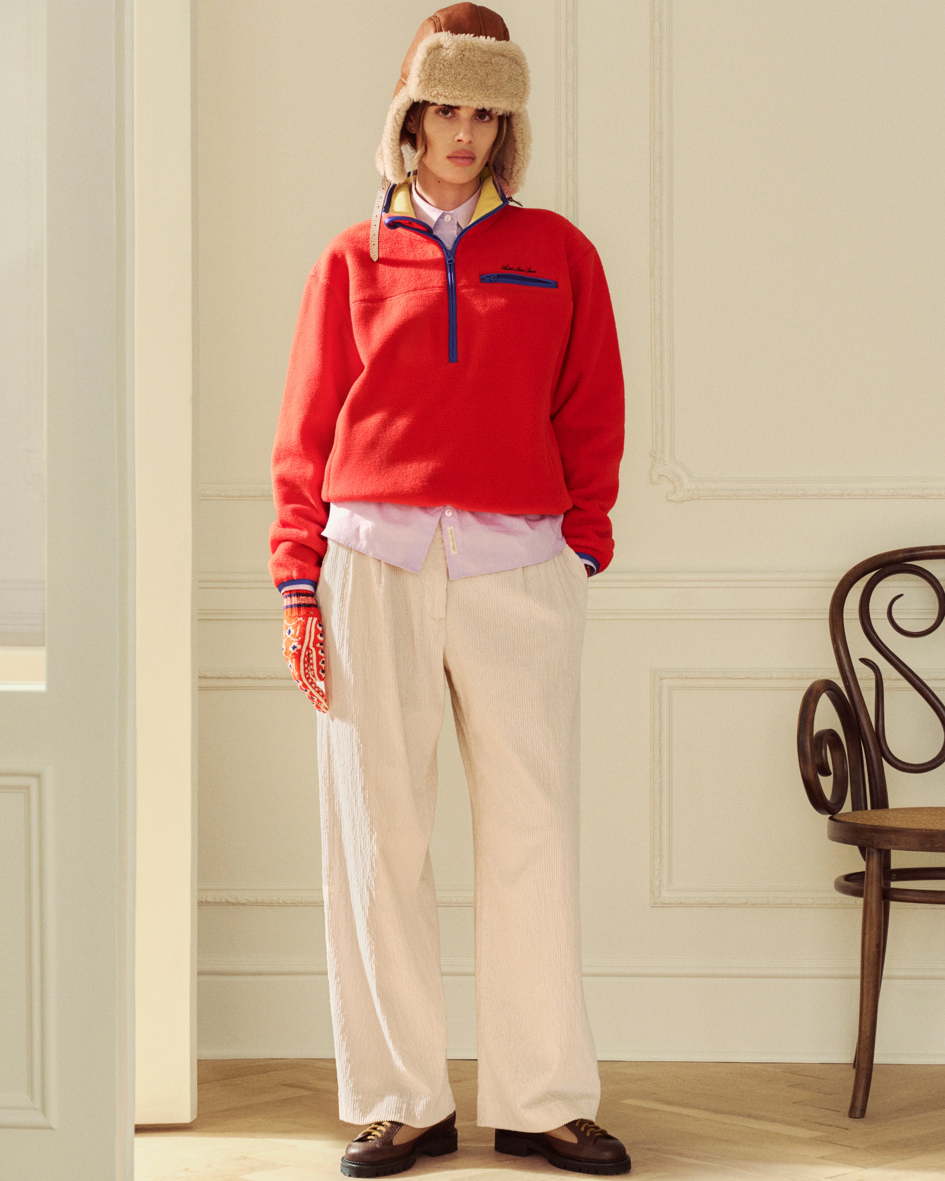 A person wearing a red Script Fleece Half Zip Pullover, light shirt, beige trousers, and a fuzzy trapper hat.