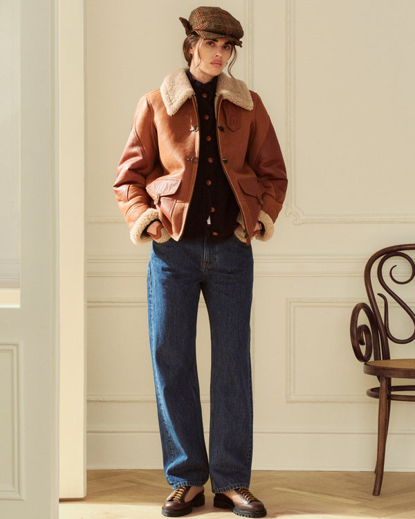 A person wearing a brown shearling-lined leather jacket, dark cardigan, flat cap, and ALD Relaxed Fit Denim jeans.