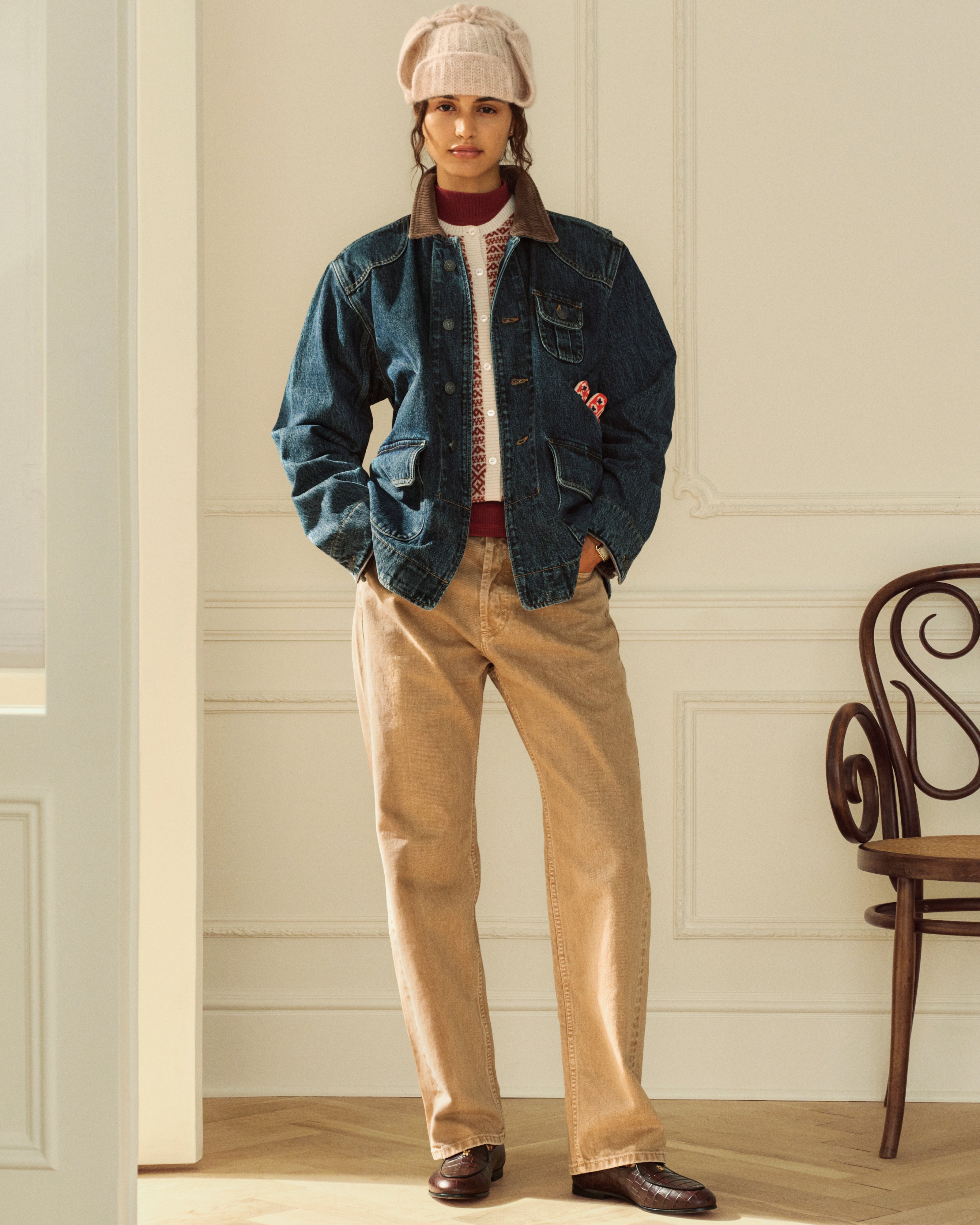 A person wears a dark Denim Chore Coat over a patterned sweater with tan trousers.