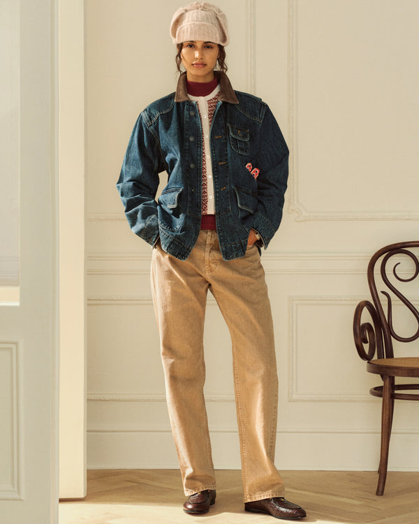 A person wears a dark Denim Chore Coat over a patterned sweater with tan trousers.