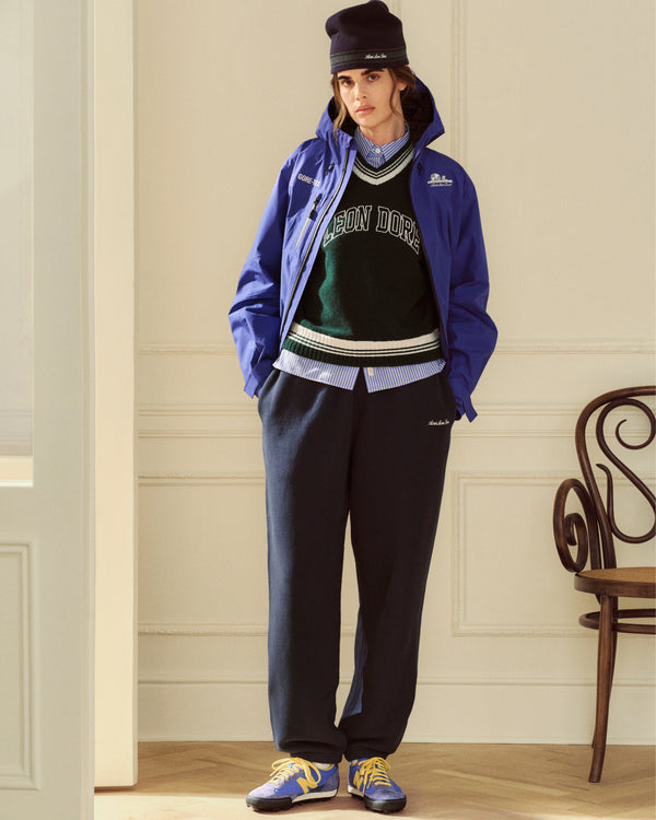 Person wearing a blue Cropped GORE-TEX Shell Jacket over a green v-neck sweater, collared shirt, trousers, beanie, sneakers.