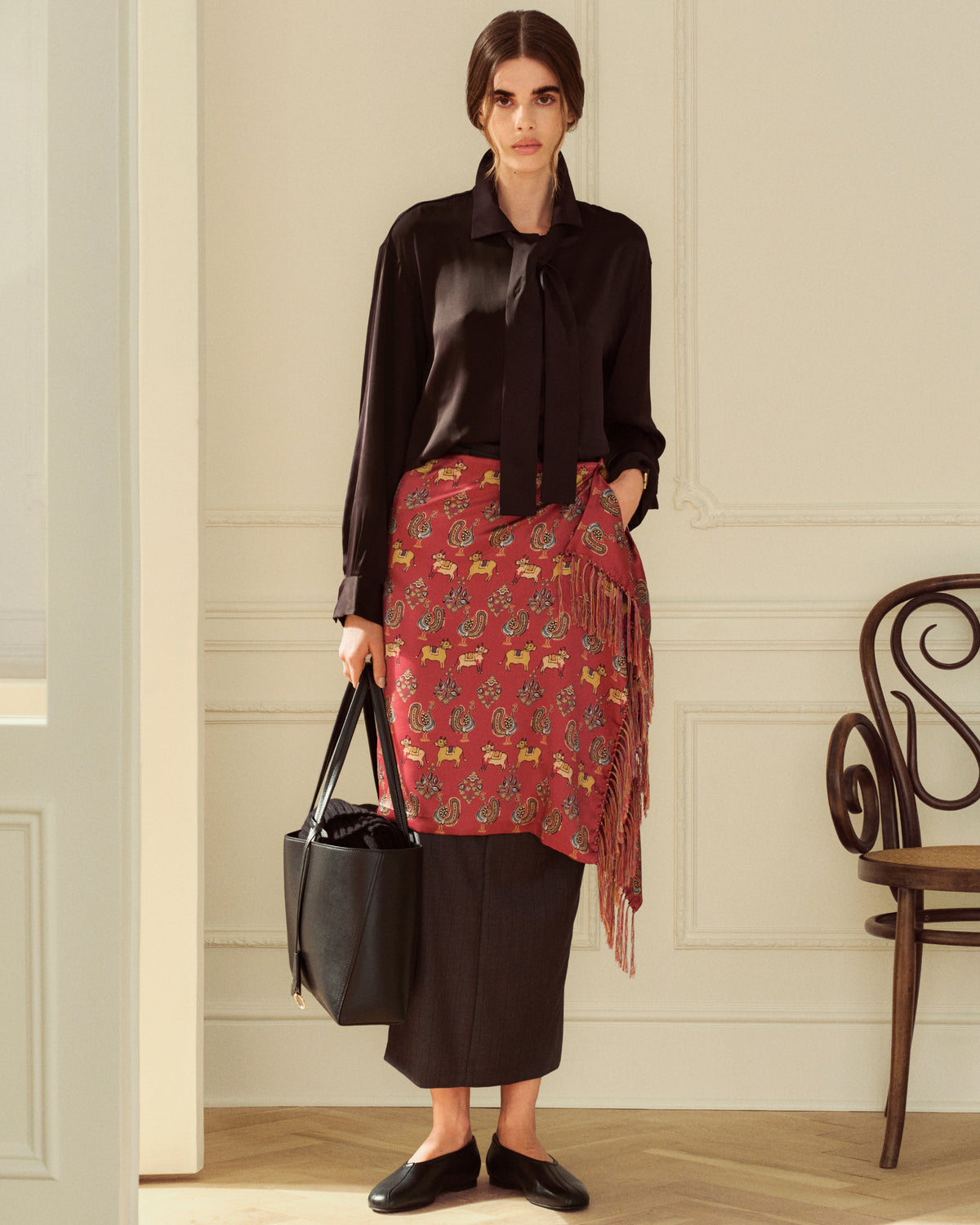 A person wearing a black blouse, a long patterned skirt, and black Leather Ballet Flat, holding a black tote bag.