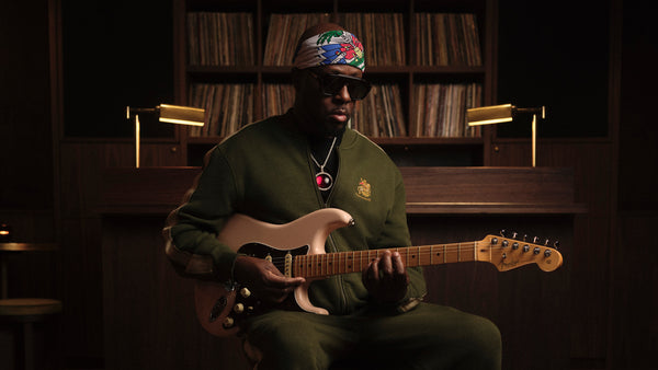 A man wearing a green tracksuit, sunglasses, and a colorful cap sits in a dimly lit room with vinyl records in the background, holding and playing a light-colored electric guitar.