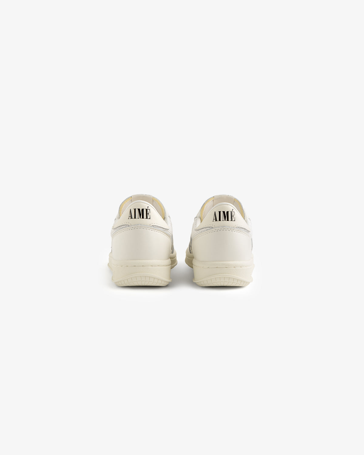 A pair of ALD / New Balance T500 Tennis Oxfords in white, seen from behind with AIMÉ in black on the heels.