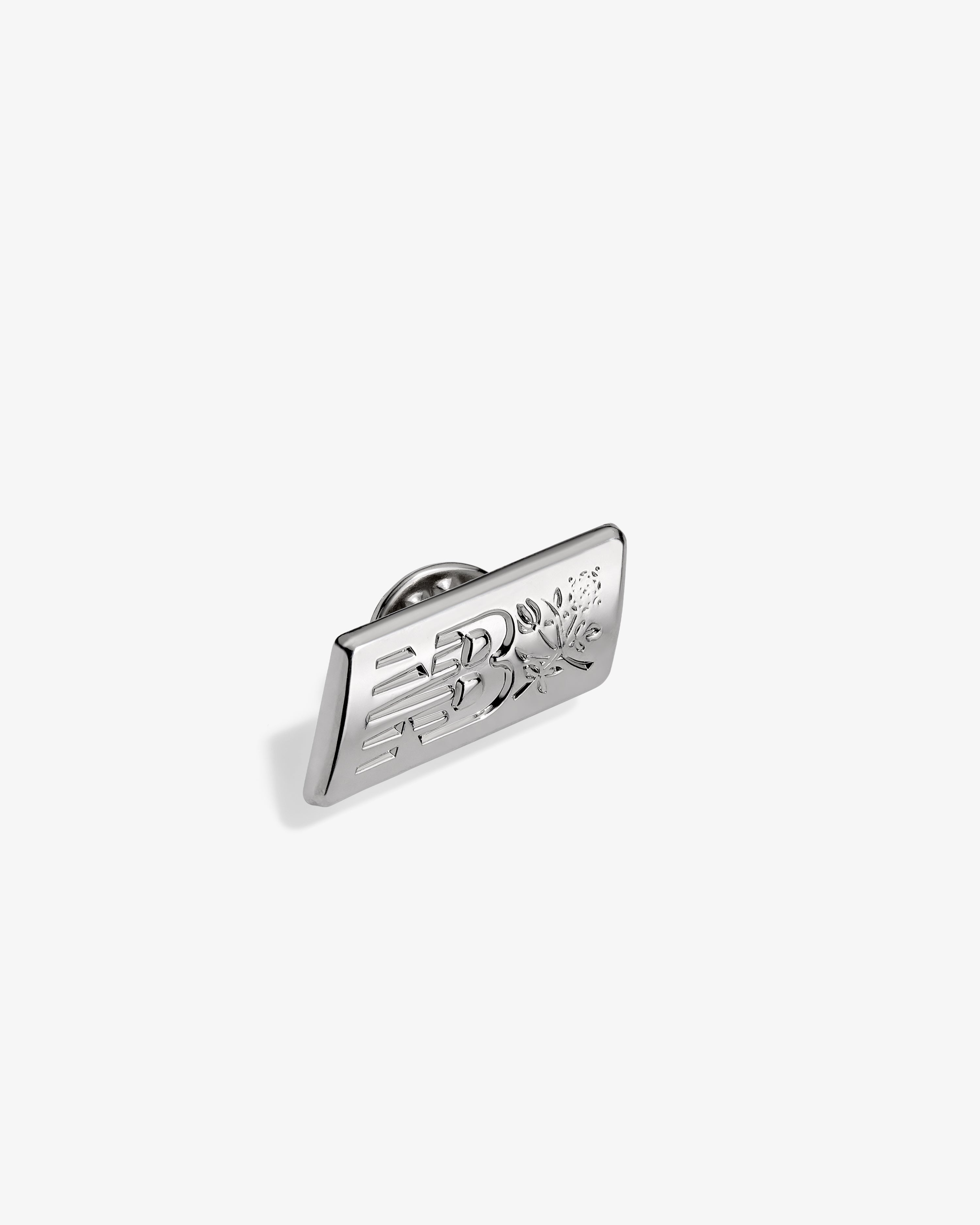 ALD / New Balance Logo Pin: rectangular silver pin with floral and geometric engraving on white background.