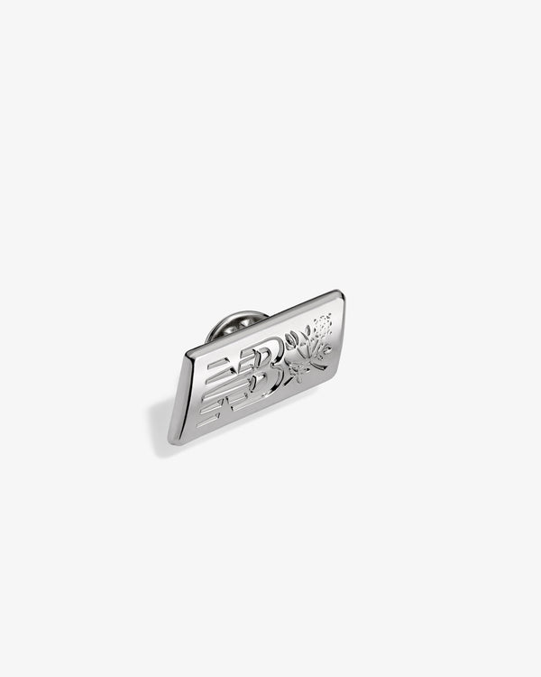 ALD / New Balance Logo Pin: rectangular silver pin with floral and geometric engraving on white background.