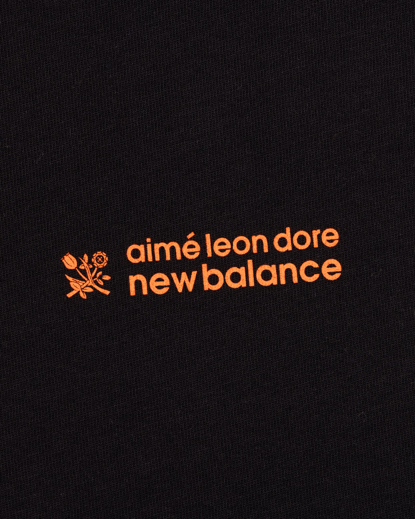 Black ALD / New Balance Running Bib Tee with orange text and a small floral graphic on black fabric.