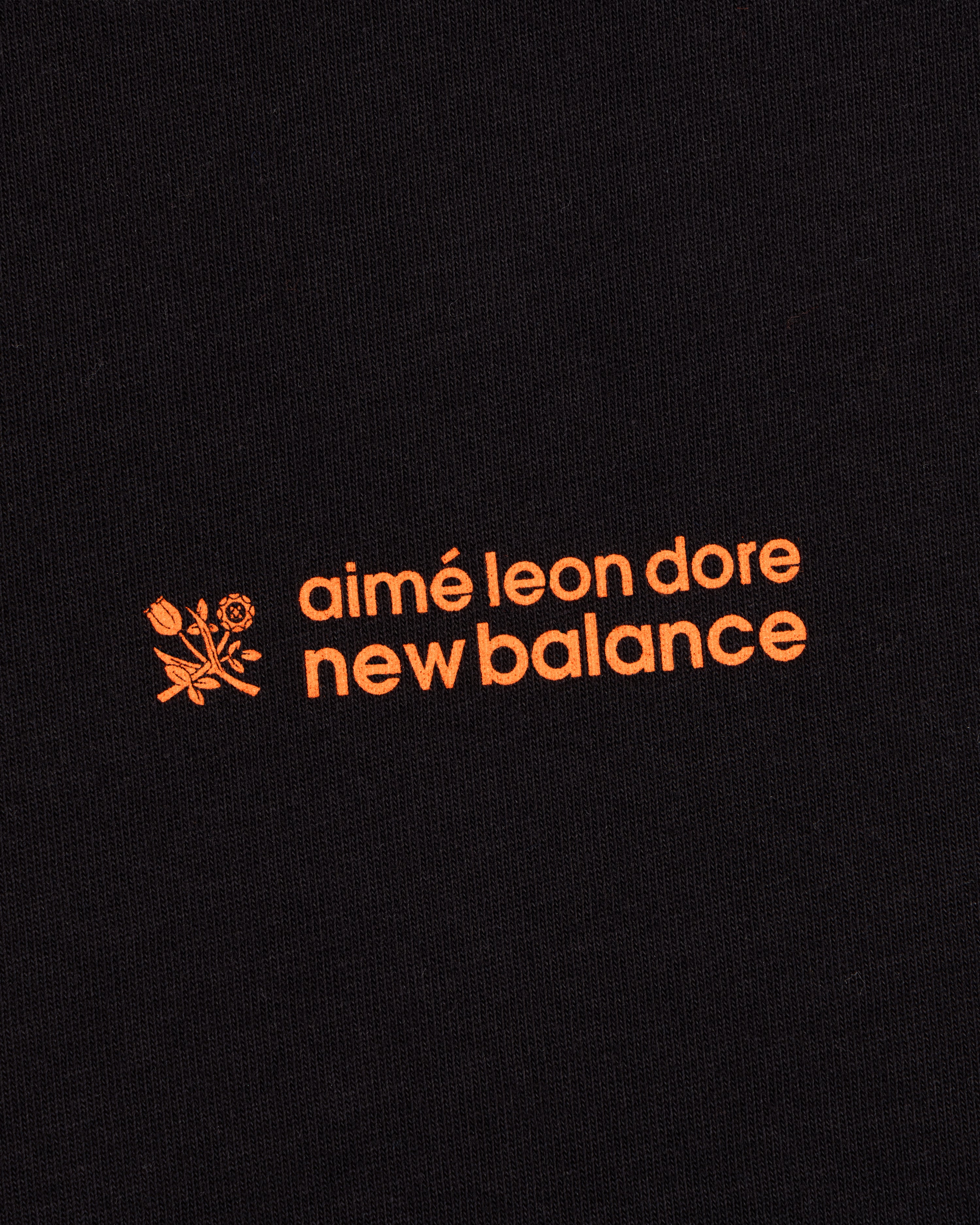 Black ALD / New Balance Running Bib Tee with orange text and a small floral graphic on black fabric.