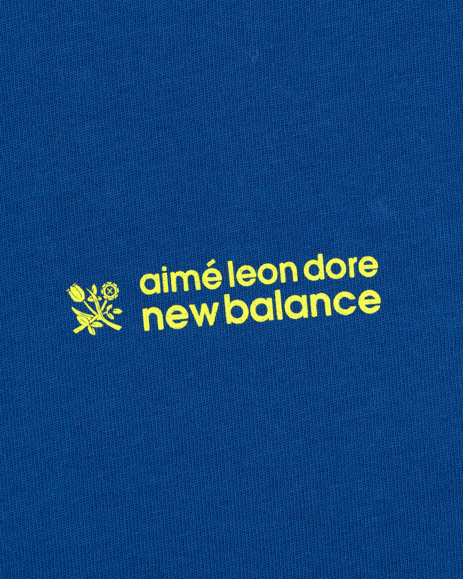 Yellow "ALD / New Balance Running Bib Tee" text and a small flower graphic on blue fabric.