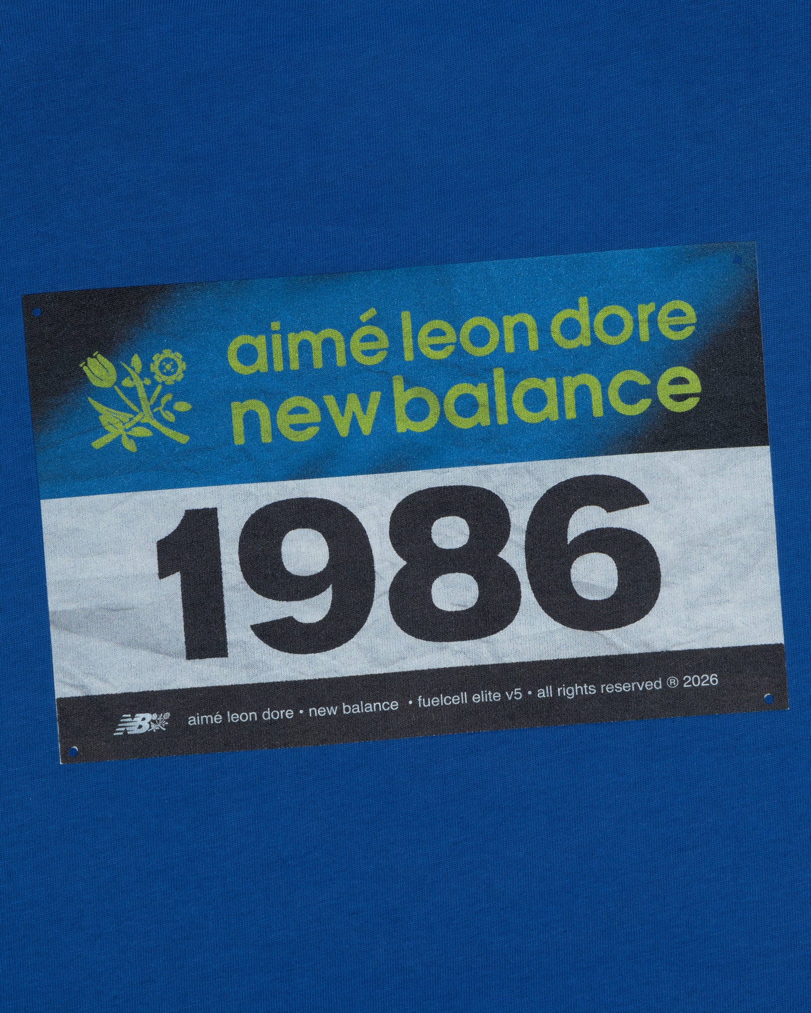 Blue background featuring the ALD / New Balance Running Bib Tee with a bold 1986 race bib graphic.