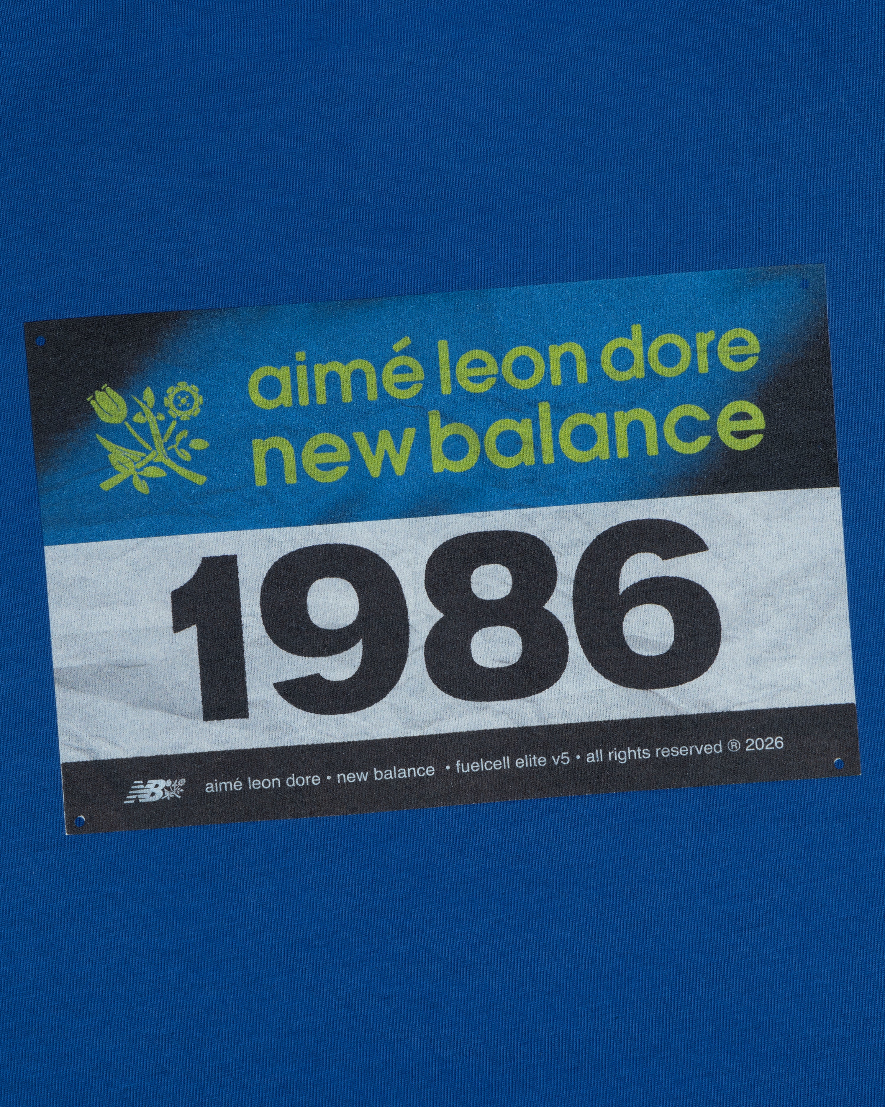 Blue background featuring the ALD / New Balance Running Bib Tee with a bold 1986 race bib graphic.