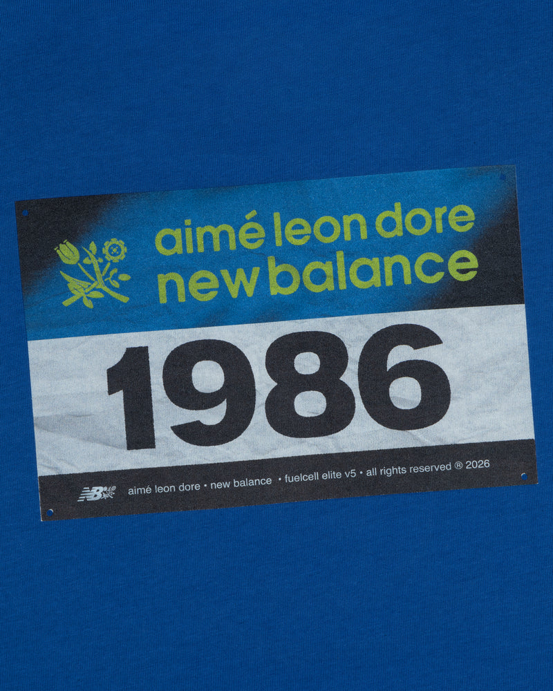 Blue background featuring the ALD / New Balance Running Bib Tee with a bold 1986 race bib graphic.