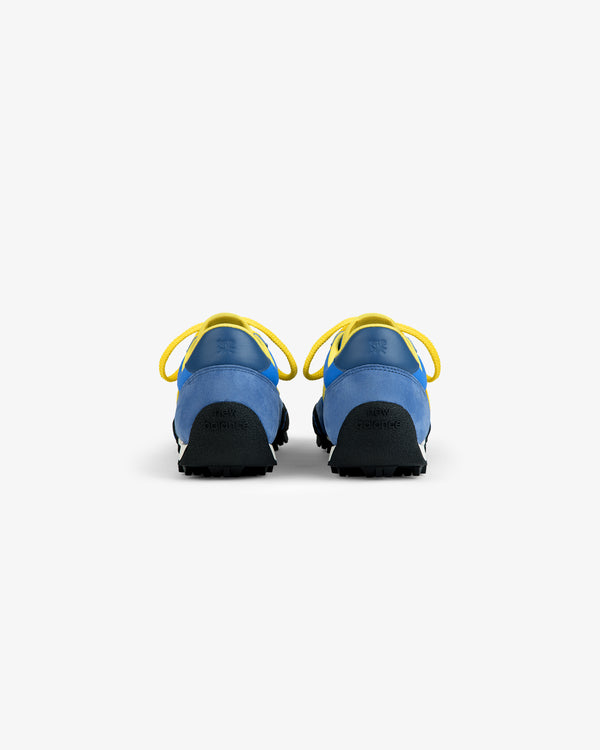 Back view of two blue ALD / New Balance Gator athletic sneakers with yellow laces and black lugged rubber soles.