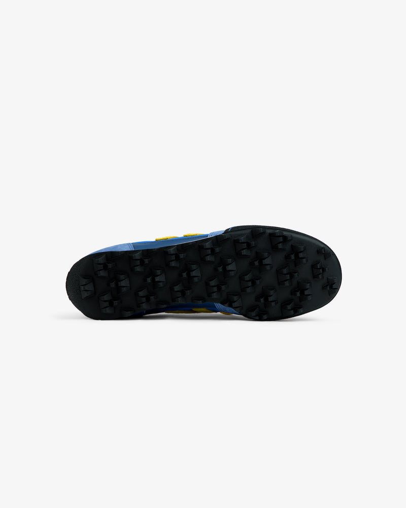 Bottom view of an ALD / New Balance Gator sneaker with a blue upper and a black, cleated rubber sole.