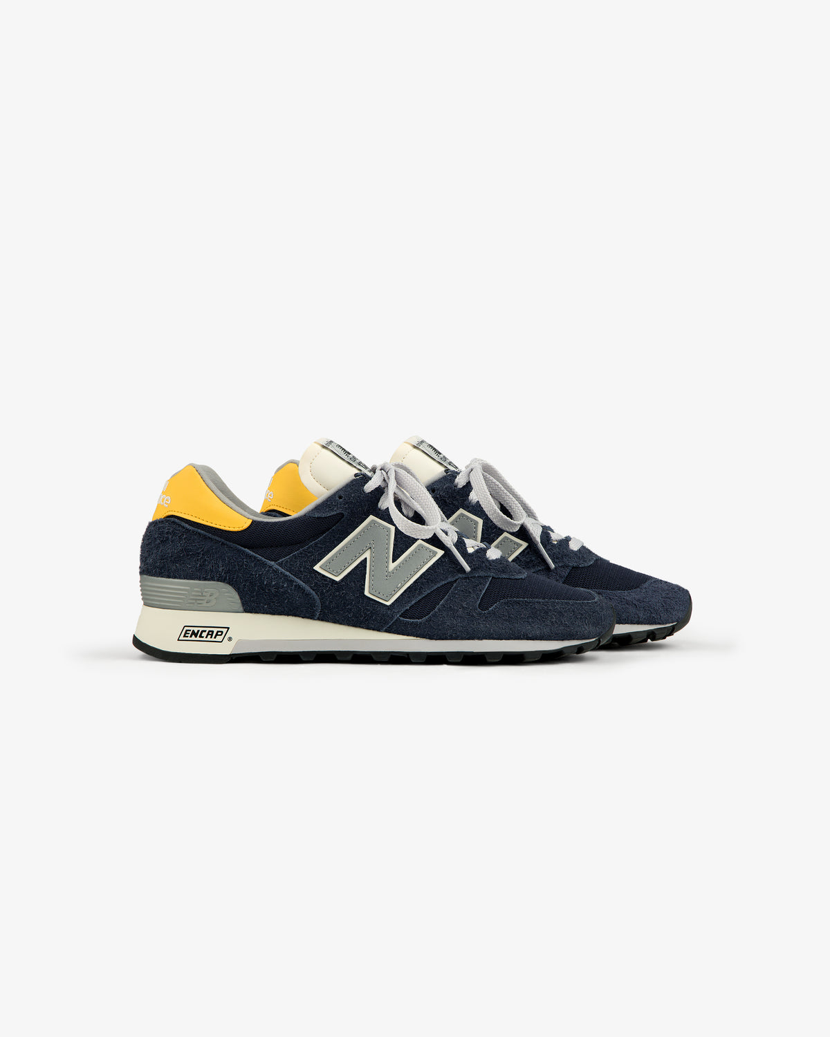 A side view of ALD / New Balance Made in USA 1300 sneakers in navy with yellow heel accents and white laces.