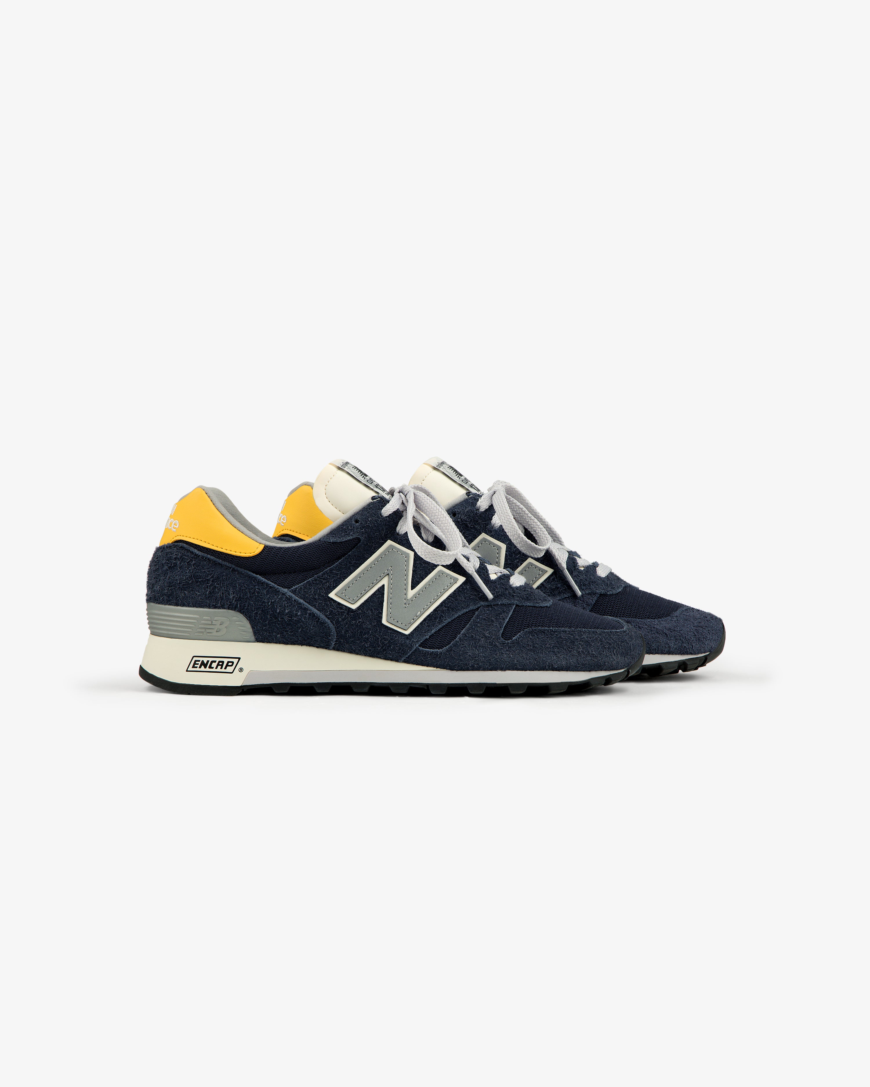 A side view of ALD / New Balance Made in USA 1300 sneakers in navy with yellow heel accents and white laces.