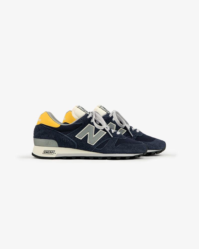 A side view of ALD / New Balance Made in USA 1300 sneakers in navy with yellow heel accents and white laces.