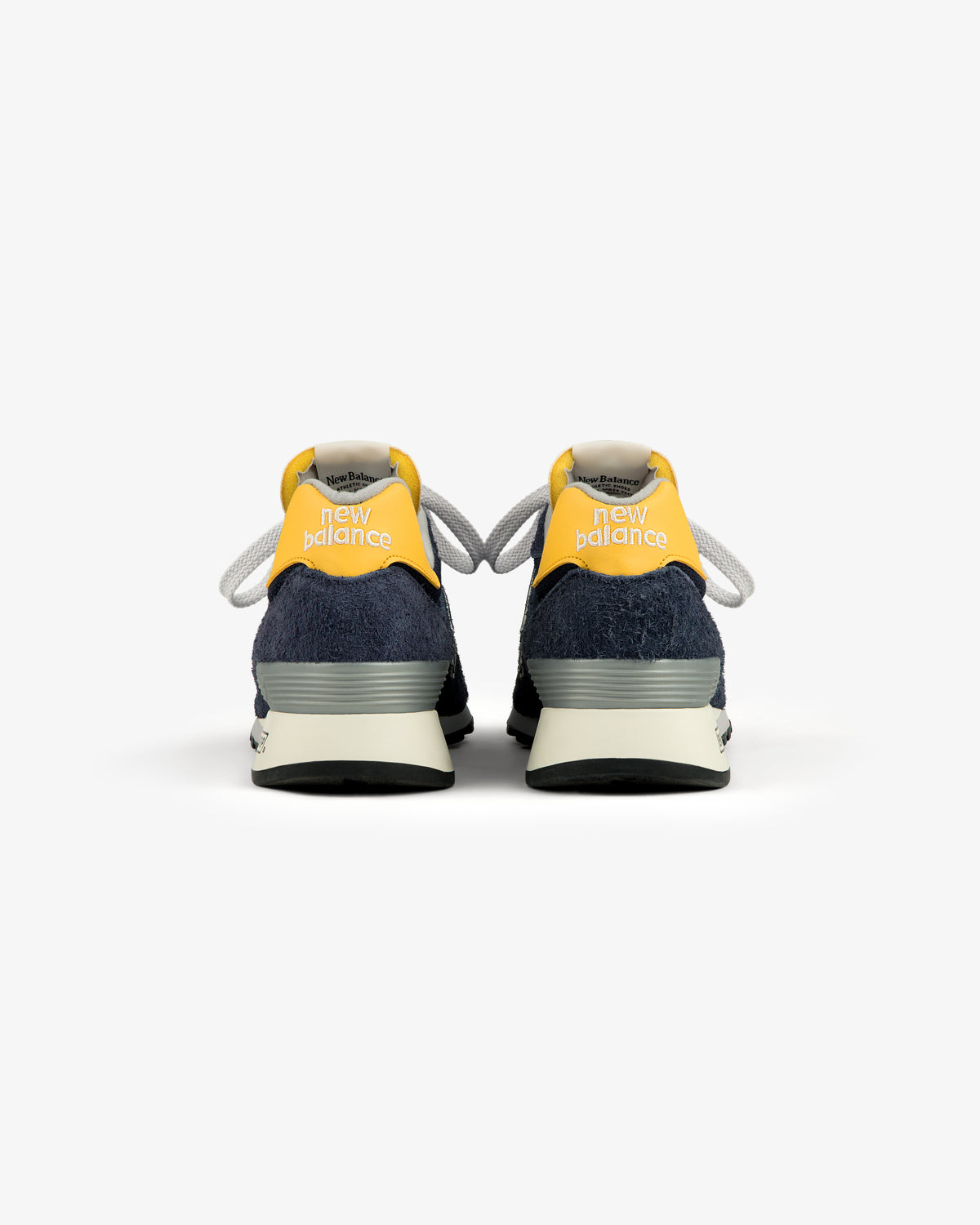 The ALD / New Balance Made in USA 1300 sneakers with yellow heels and navy blue suede accents, shown from the back.
