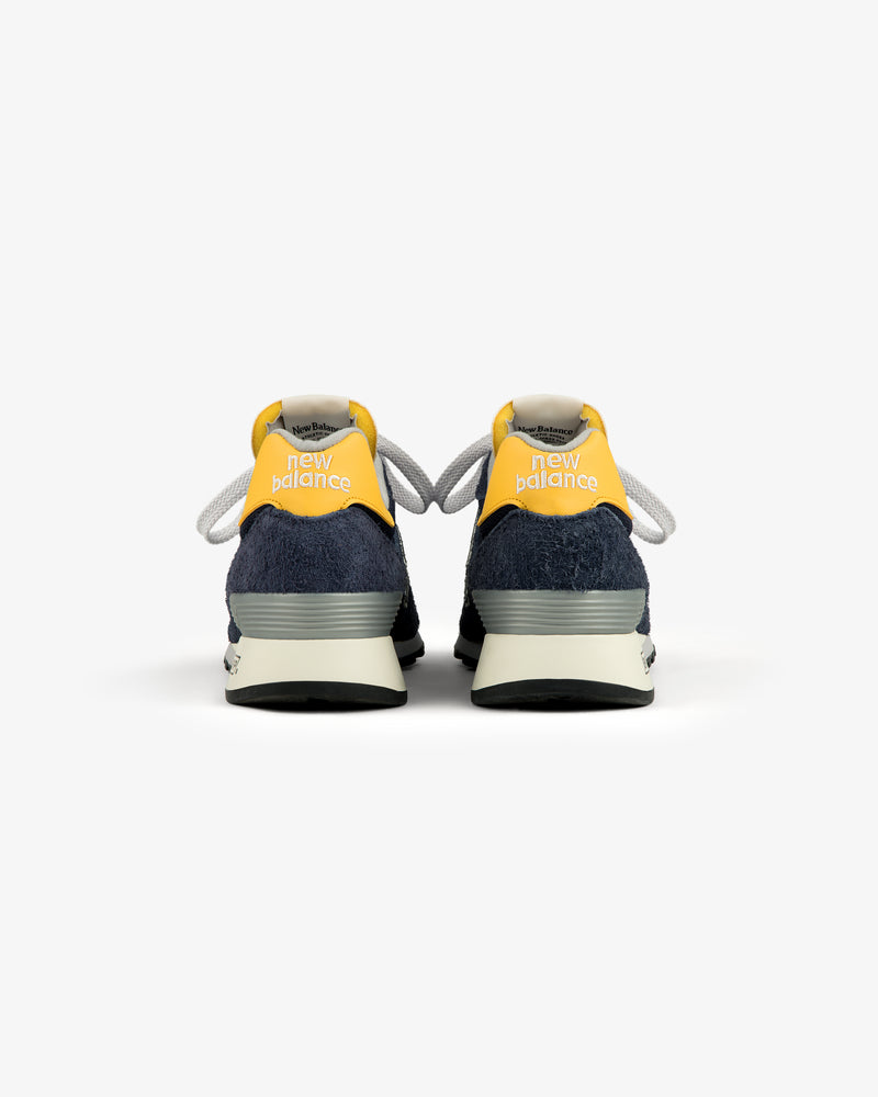 The ALD / New Balance Made in USA 1300 sneakers with yellow heels and navy blue suede accents, shown from the back.
