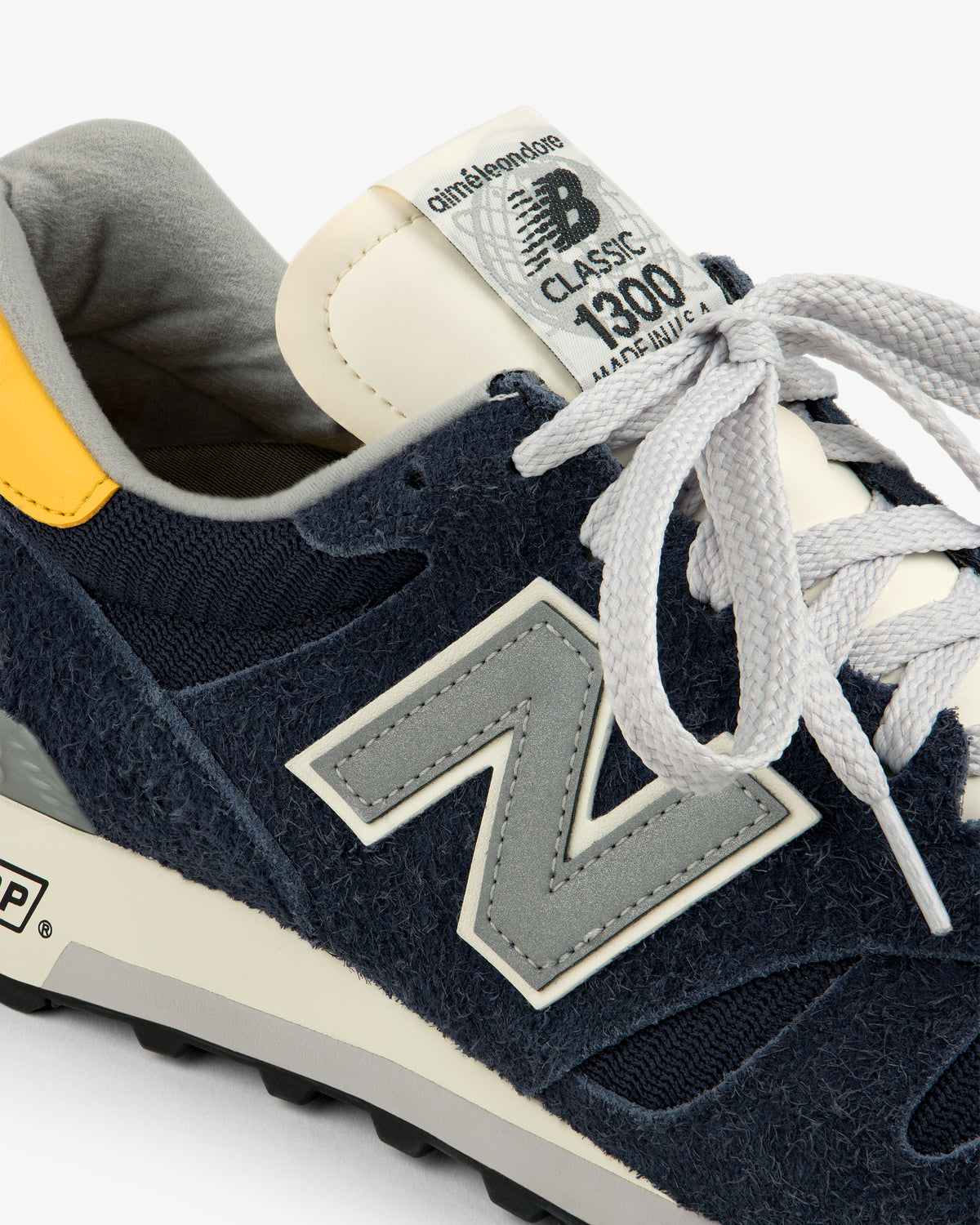 Close-up of an ALD / New Balance Made in USA 1300 sneaker in navy blue with grey laces and a yellow heel accent.